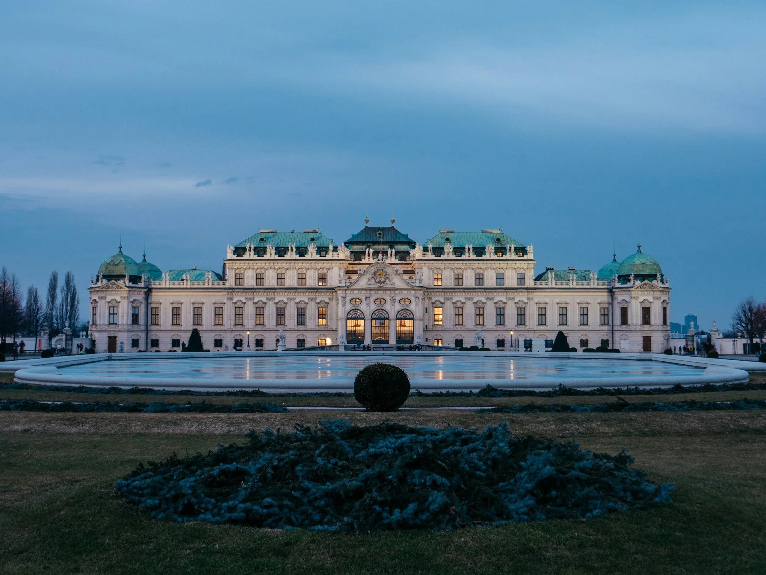 The Schönbrunn Palace in Vienna, Austria, illuminated at dusk with a reflecting pool and landscaped garden in the foreground.