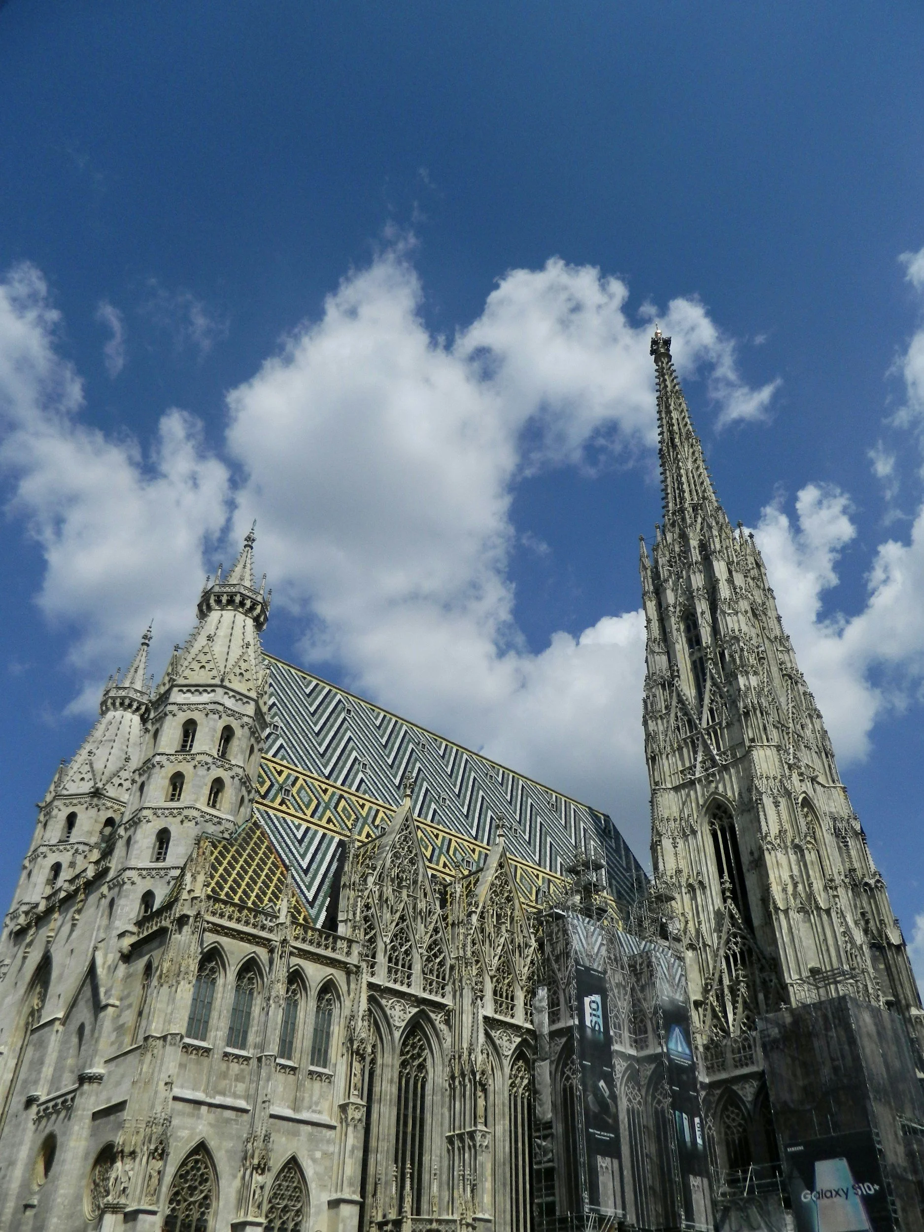 A Gothic cathedral with intricate stone details and a colorful tiled roof, set against a blue sky with white clouds.