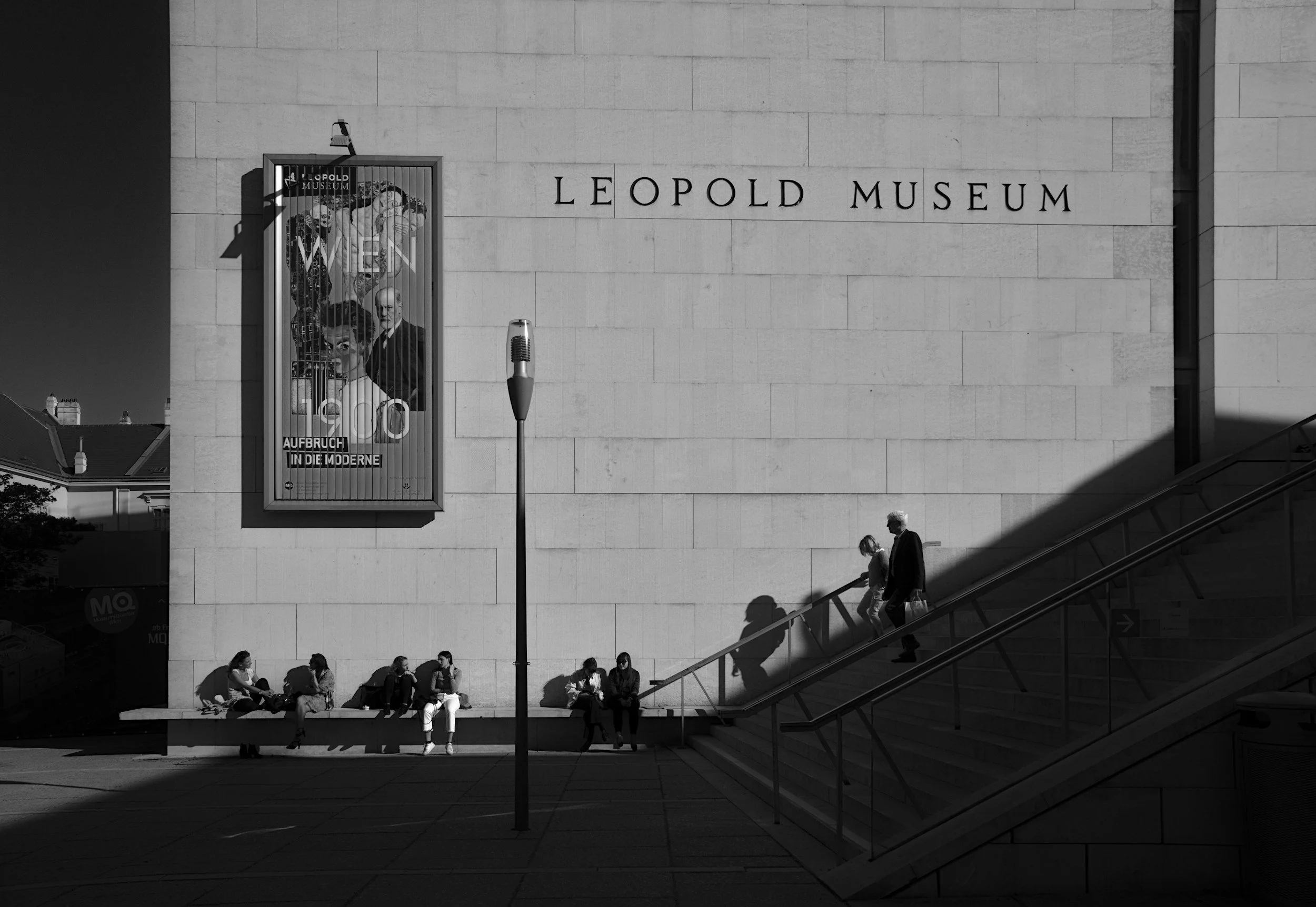 People sitting on benches and walking up stairs outside the Leopold Museum in Berlin, with the museum's name on the building and a poster advertisement on the wall.
