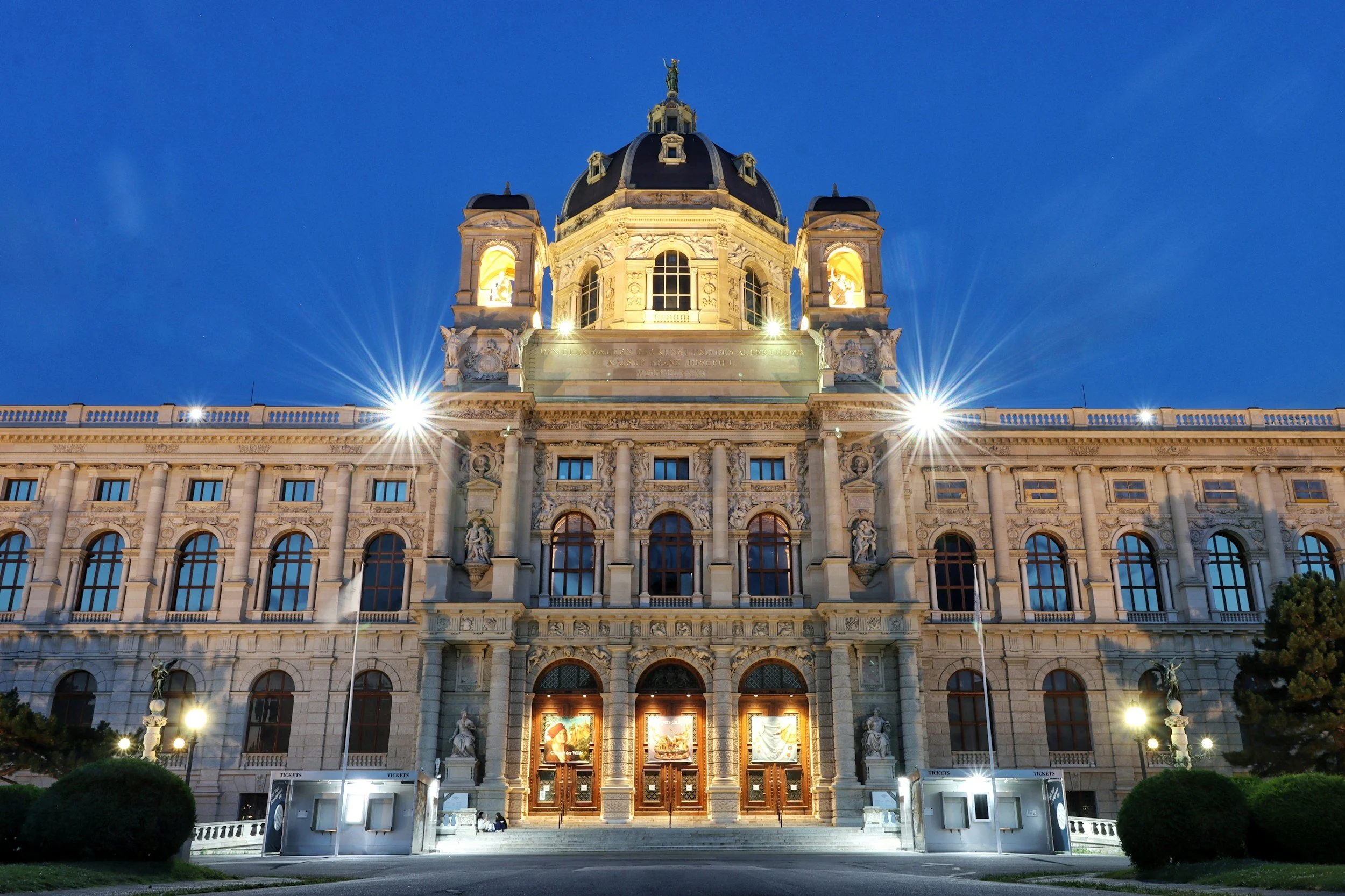 Night view of a historic ornate building with a dome and statues, illuminated with exterior lighting, against a dark blue sky.
