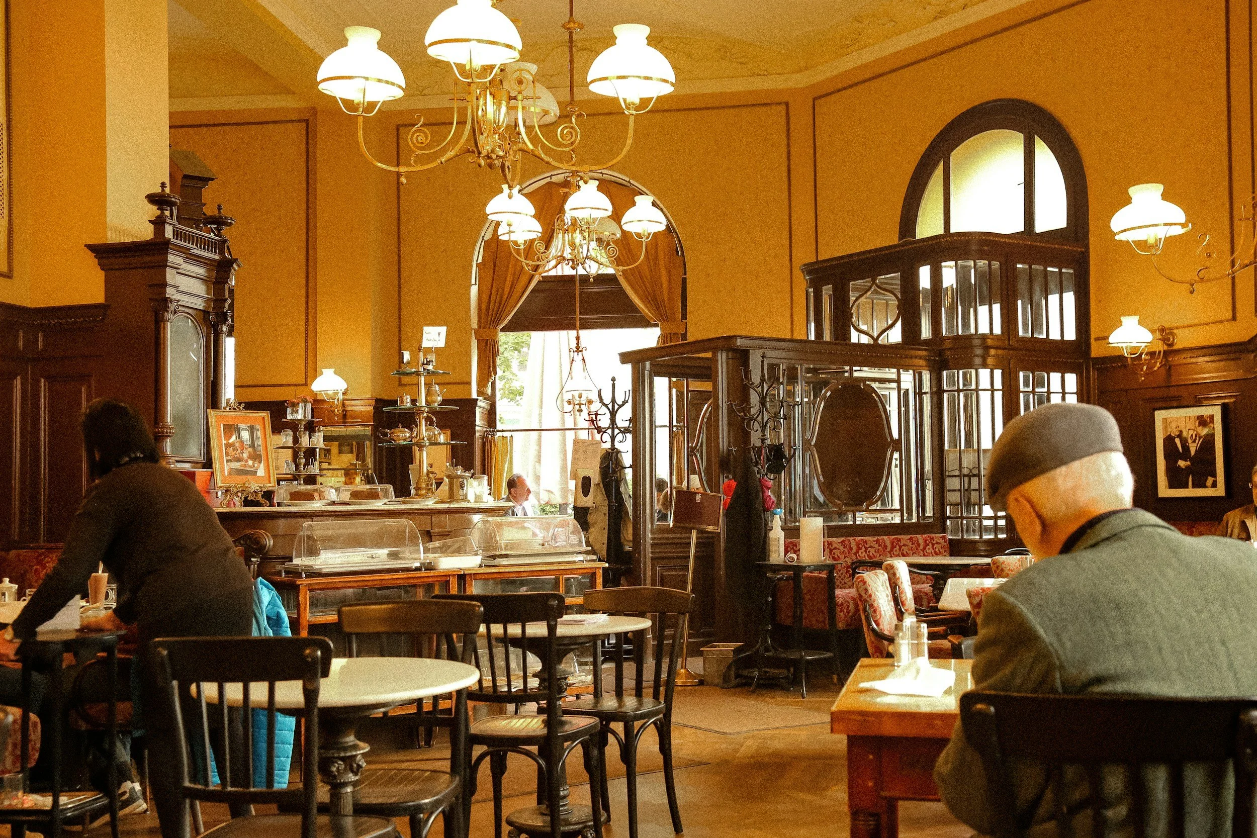 Interior of a cozy, vintage-style restaurant with warm lighting, wooden furniture, a chandelier, and a large window with curtains. Several customers are dining, including an elderly man with a beret and glasses, and a woman in dark clothing.