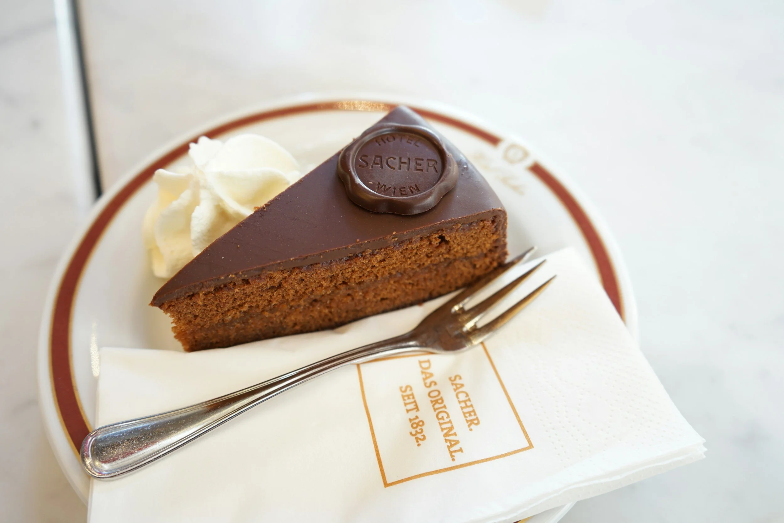 A slice of chocolate cake with a chocolate glaze, topped with a chocolate emblem, served on a white plate with a dollop of whipped cream, a fork, and a napkin.