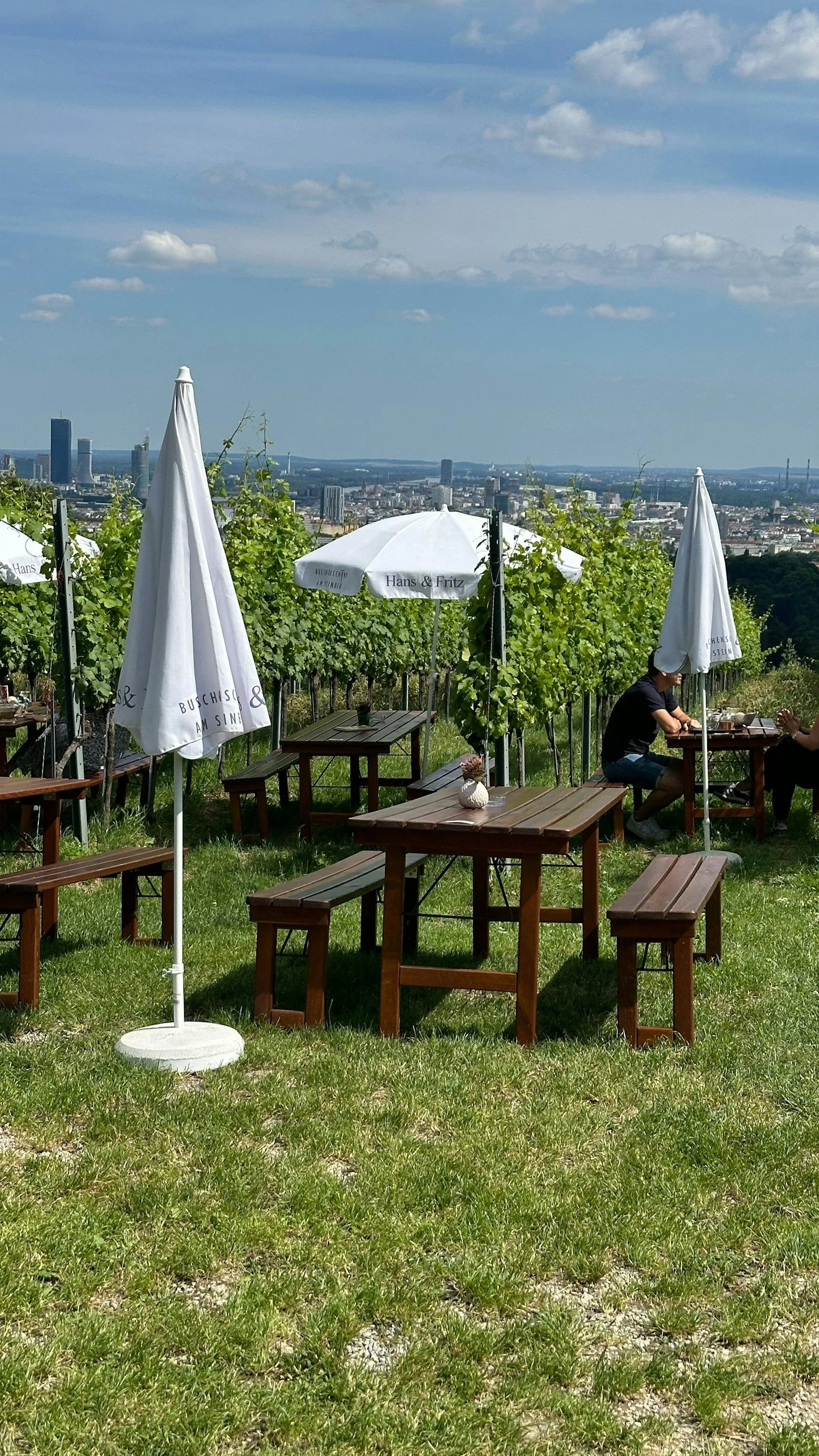 Outdoor vineyard restaurant with wooden tables, benches, and white umbrellas, set on a grassy hill overlooking a city skyline with tall buildings under a partly cloudy sky.