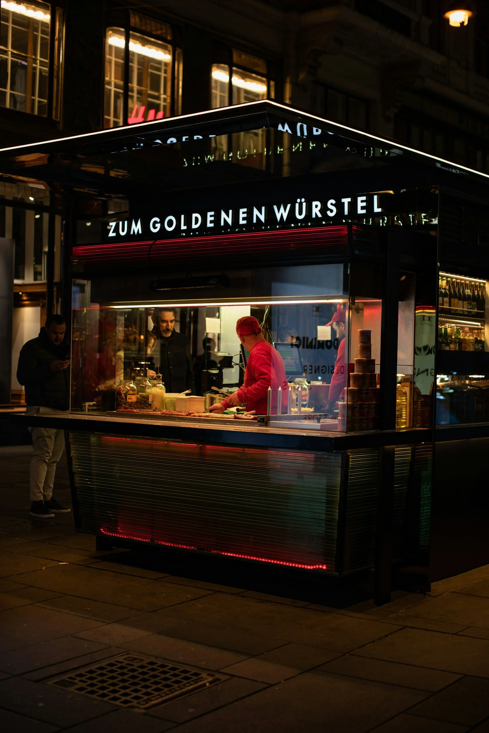 Nighttime street scene with a food stand named 'Zum Goldenen Würstel' illuminated by neon lights. The stand has a worker in a red uniform preparing food, and a customer standing nearby.