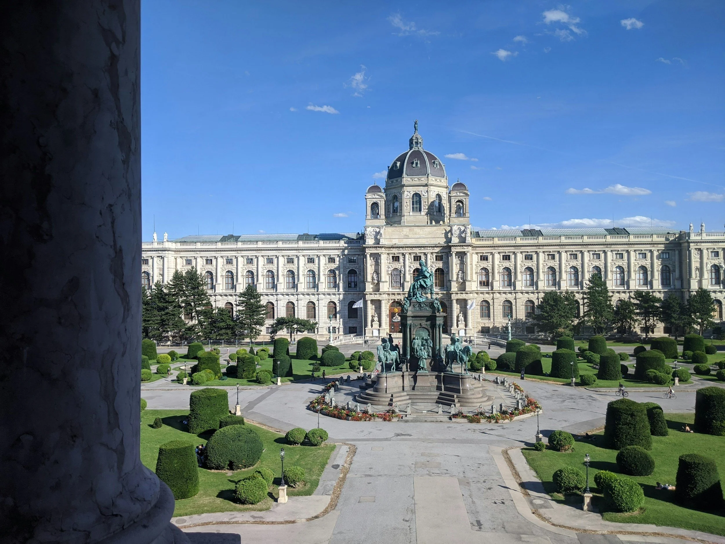 A grand historic building with a large dome, ornate stonework, and numerous windows, viewed from a garden with trimmed bushes and a statue of a man on horseback at the center.