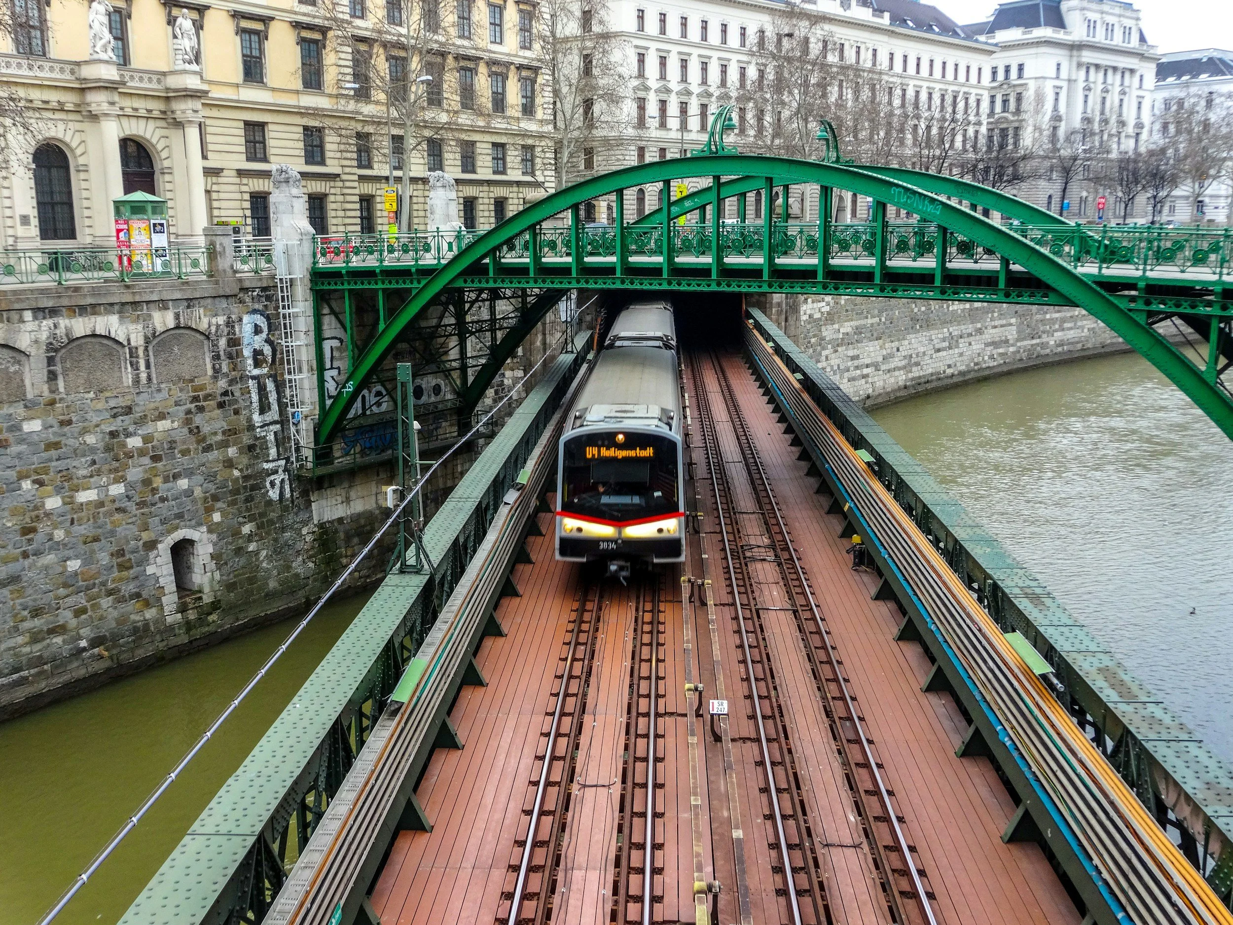 A train traveling on a bridge over a river, with a green arch above and old buildings in the background.