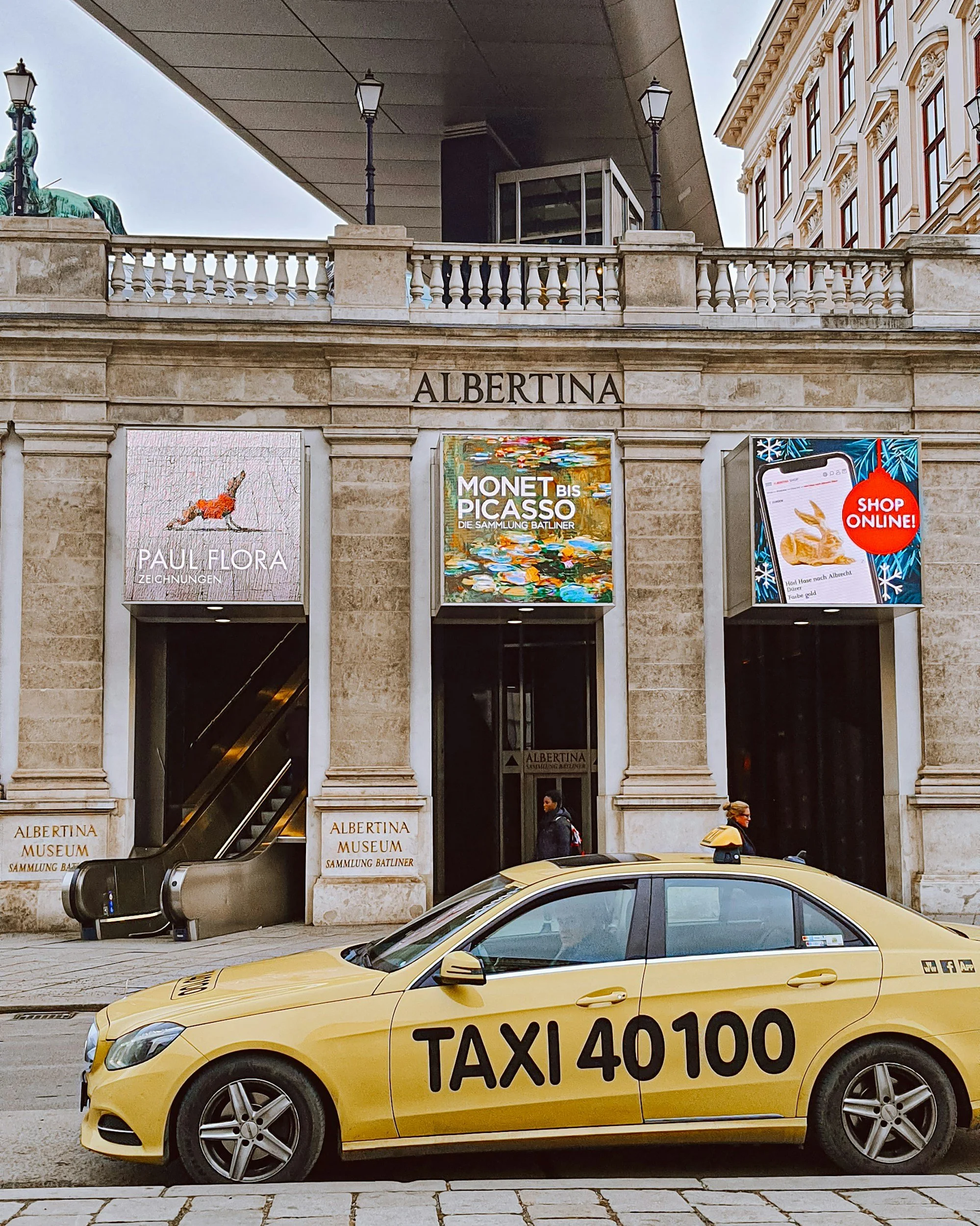 Yellow taxi cab parked on the street in front of Albertina Museum in Vienna, Austria, with digital posters and historical building architecture visible.