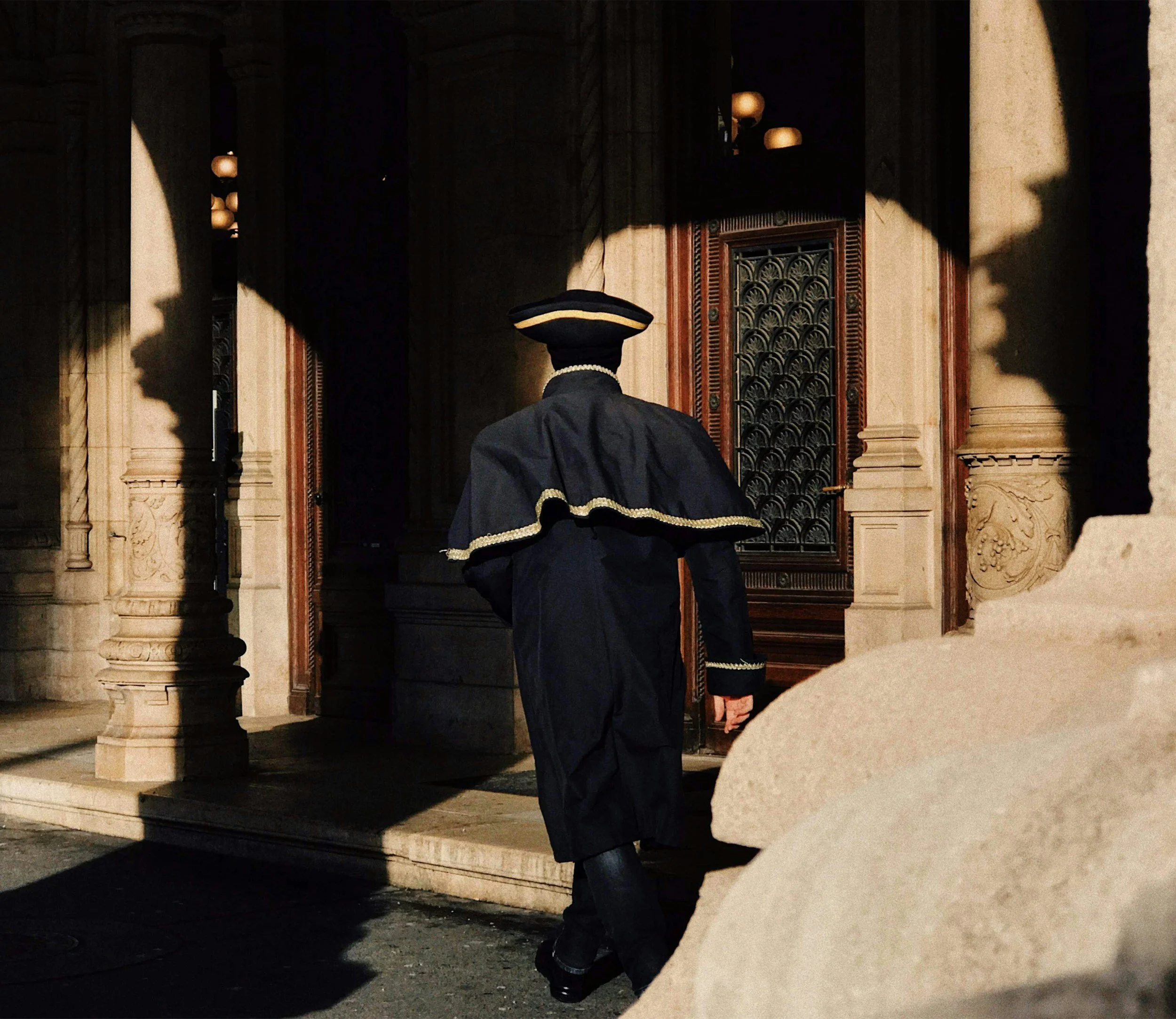 A person in historical attire walking into a building with ornate wooden doors and stone columns, shadowed by sunlight.