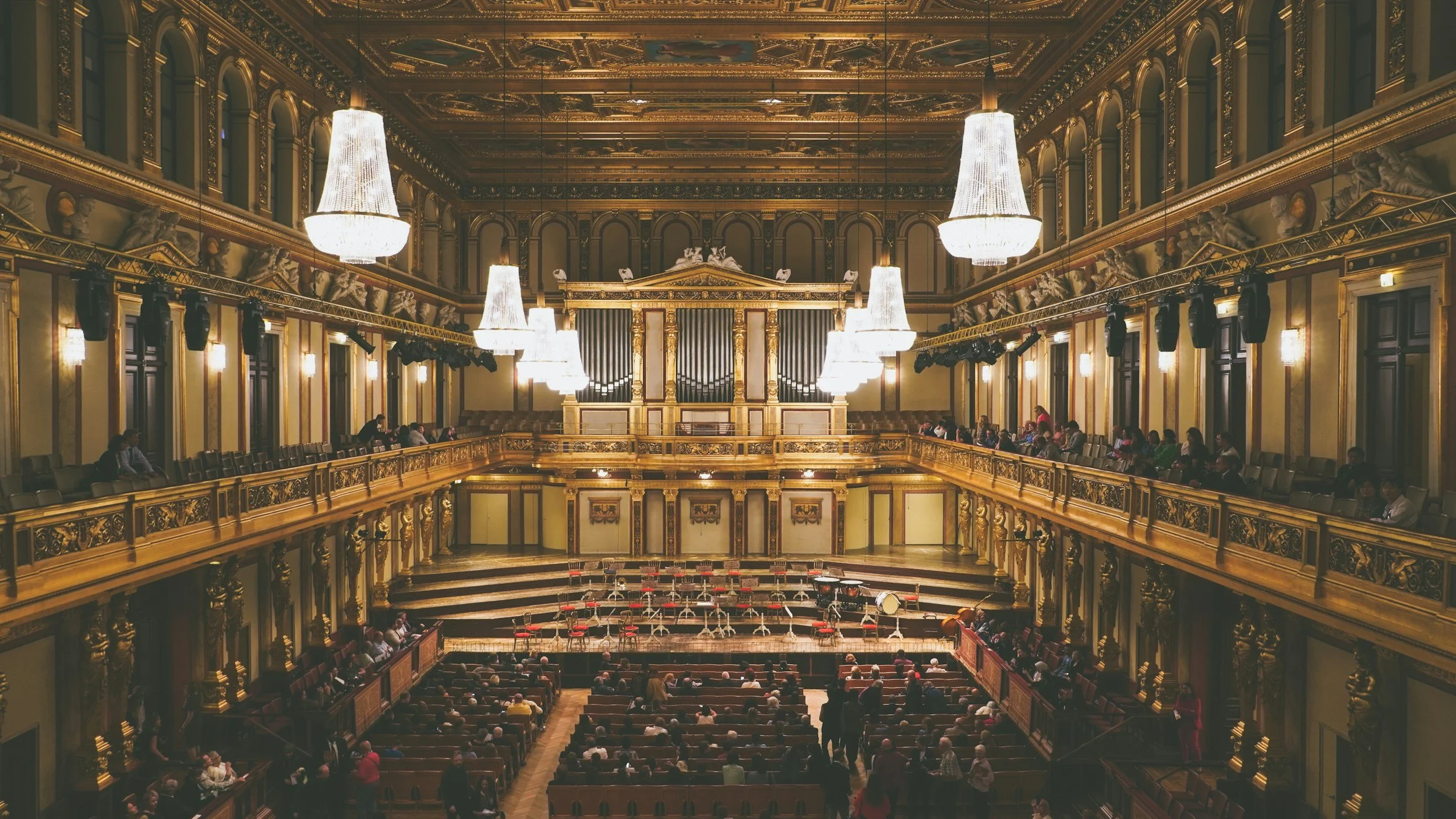 Interior of a grand concert hall with ornate gold decorations, large chandeliers, and an empty stage set up for an orchestra, with audience seated facing the stage.