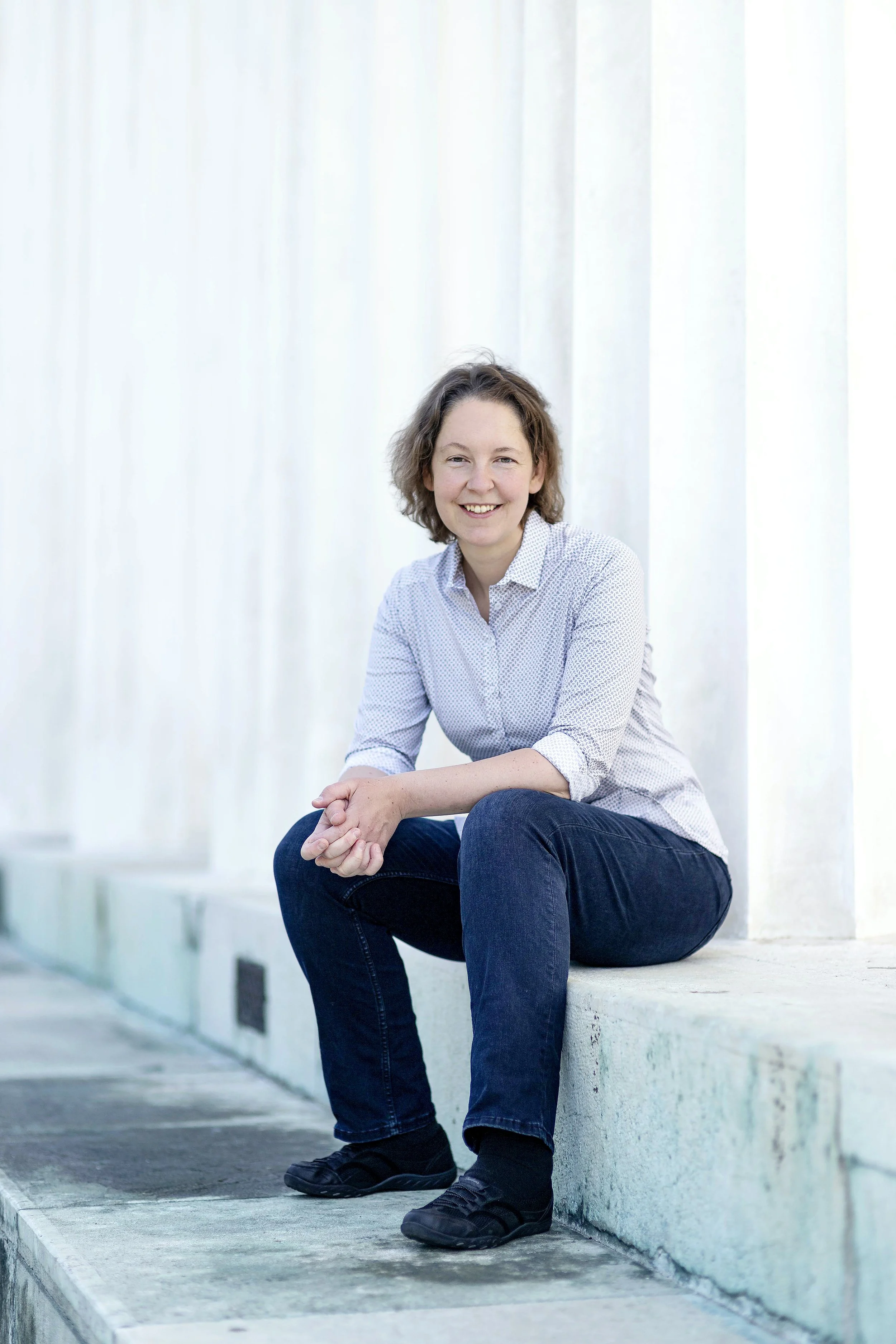 Woman sitting outdoors on concrete steps, smiling, wearing a light-colored button-up shirt and dark jeans, with a white background.