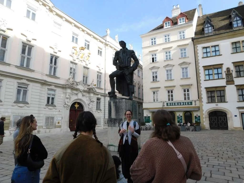A group of people gathered around a woman in front of a statue of a man sitting on a stool in a European city square, with historic buildings and a restaurant named 'Bieradies' in the background.
