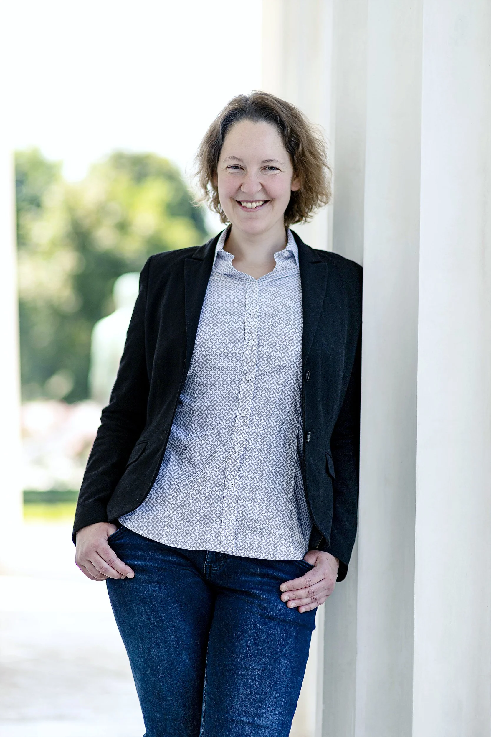 A woman with short curly hair smiling and leaning against a white pillar outdoors, with green trees in the background.
