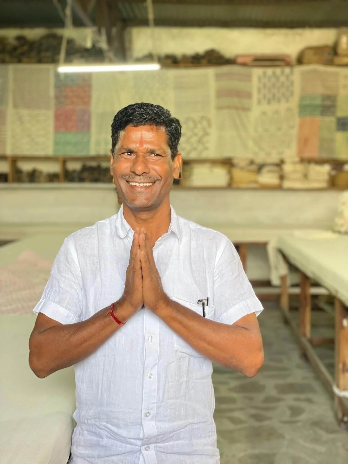 A man smiling with hands pressed together in a prayer gesture, standing in a room with colorful textiles and folded fabric in the background.