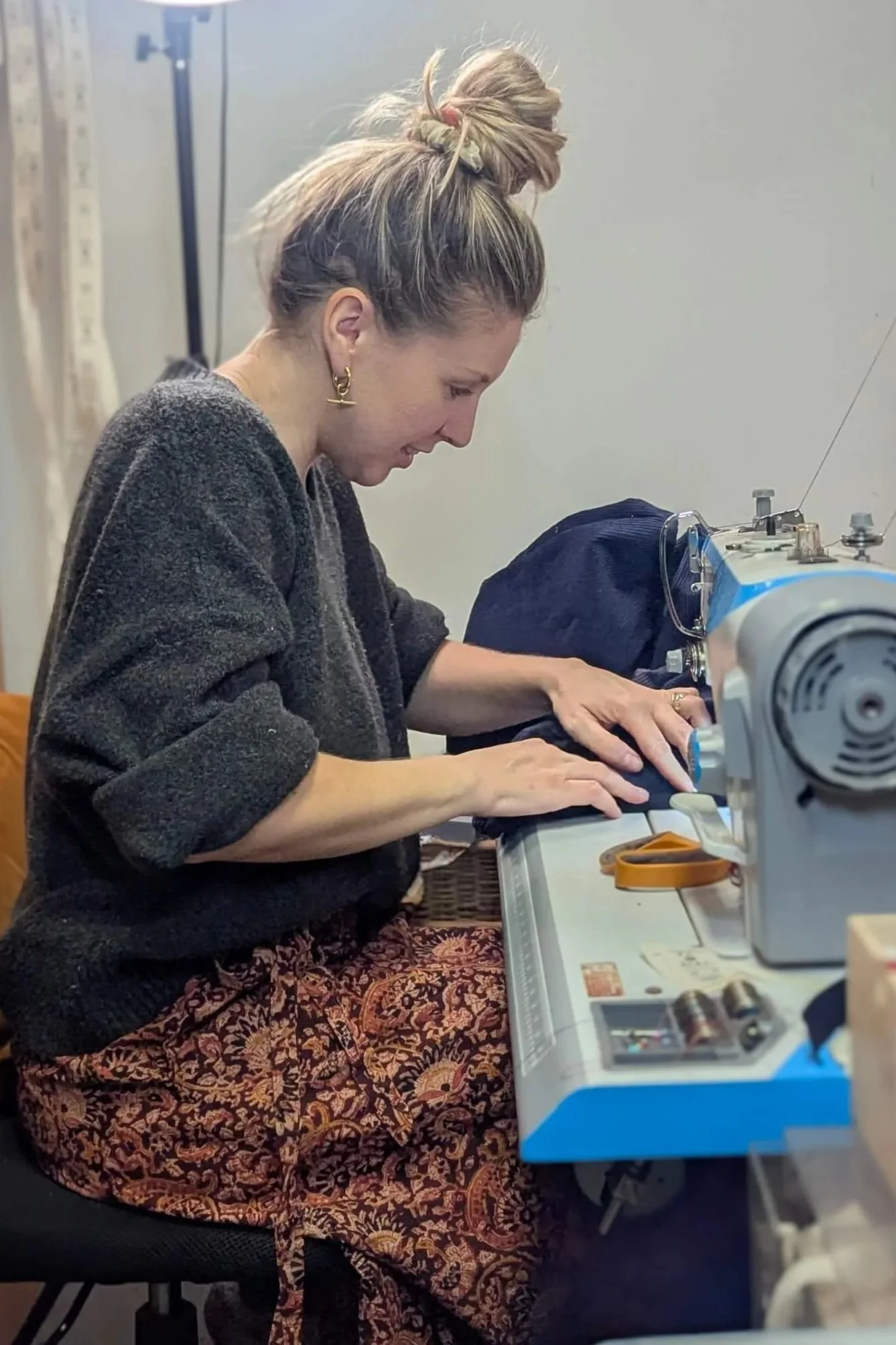 Woman sewing on a sewing machine with a patterned skirt and earrings, focusing on her work.