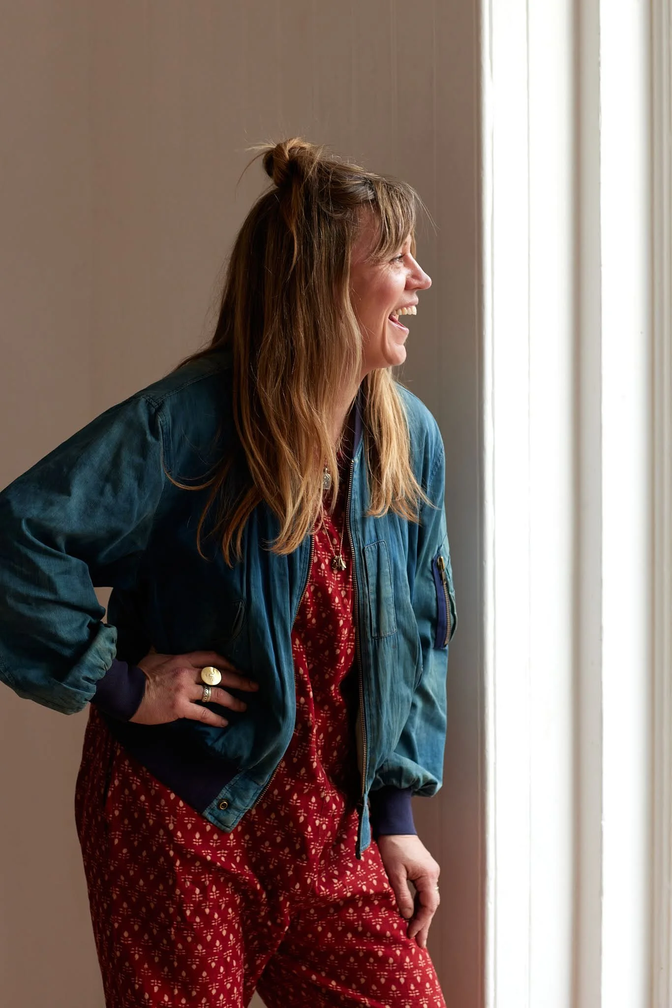 A woman with long wavy hair, smiling and looking to the right, standing near a window with curtains. She is wearing a blue jacket and a red dress with a white pattern, with her left arm akimbo and hand resting on her hip.