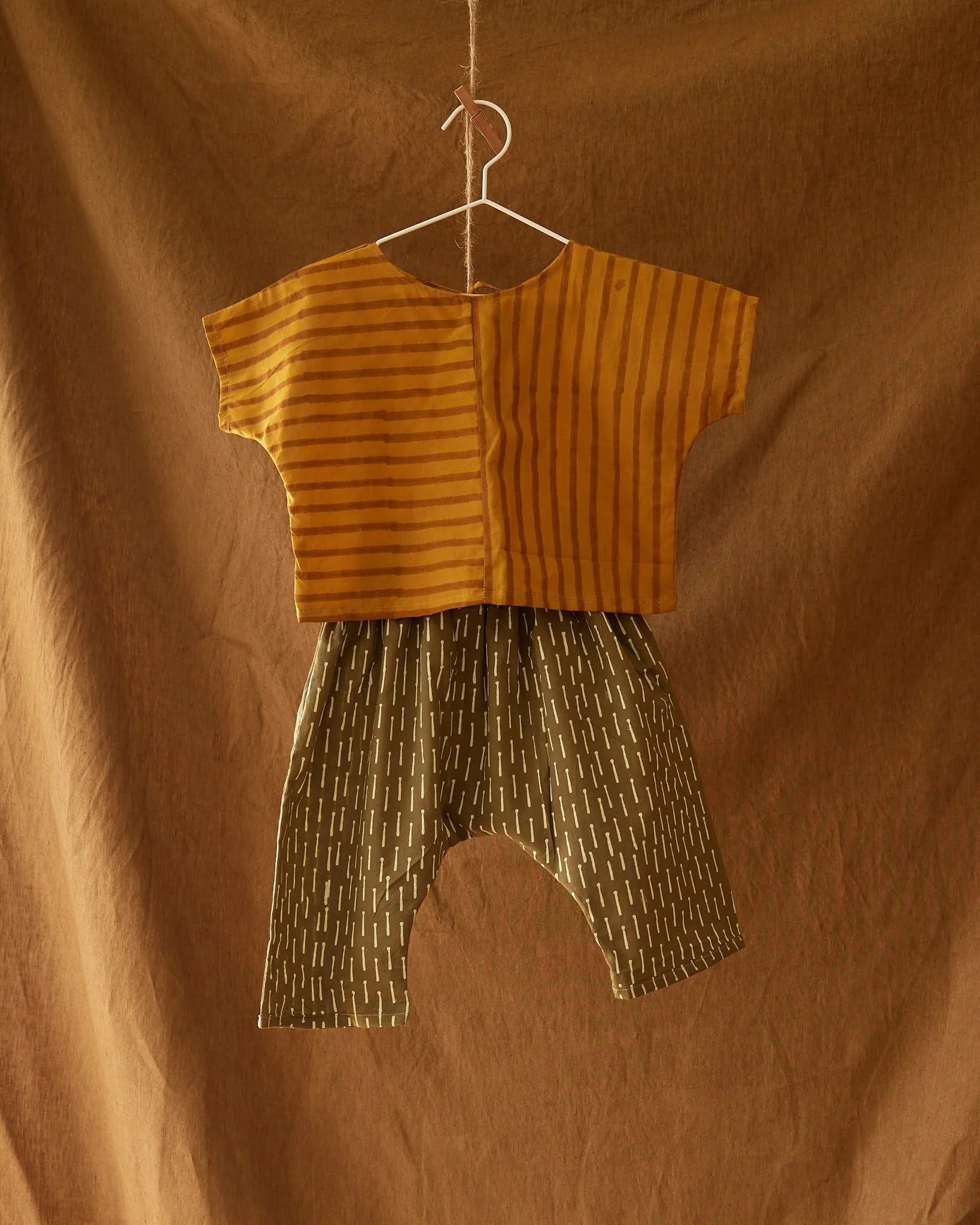 A short-sleeve yellow top with horizontal darker yellow stripes hanging on a white hanger against a brown textured background, with patterned pants hanging beneath.