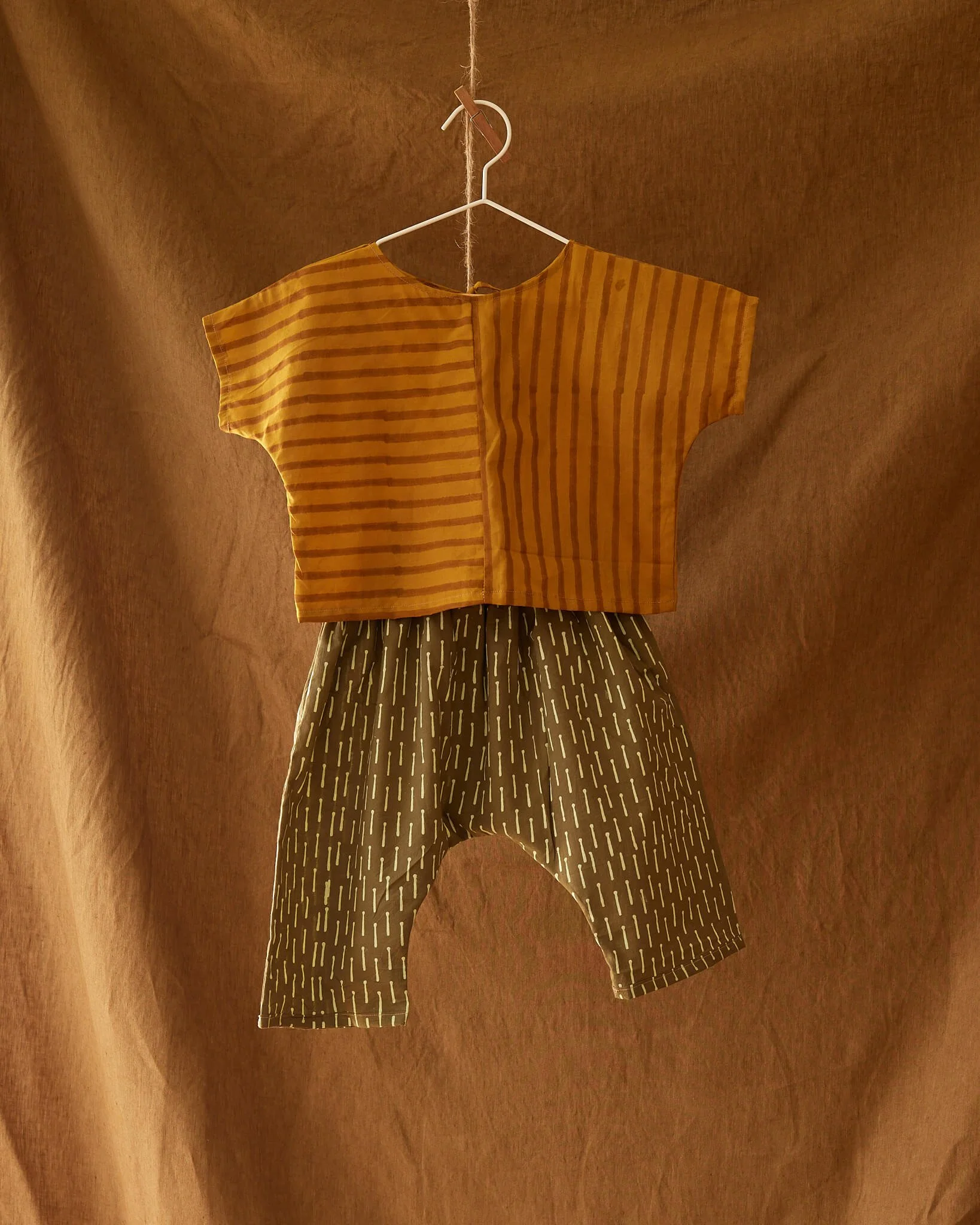 A short-sleeve yellow top with horizontal darker yellow stripes hanging on a white hanger against a brown textured background, with patterned pants hanging beneath.