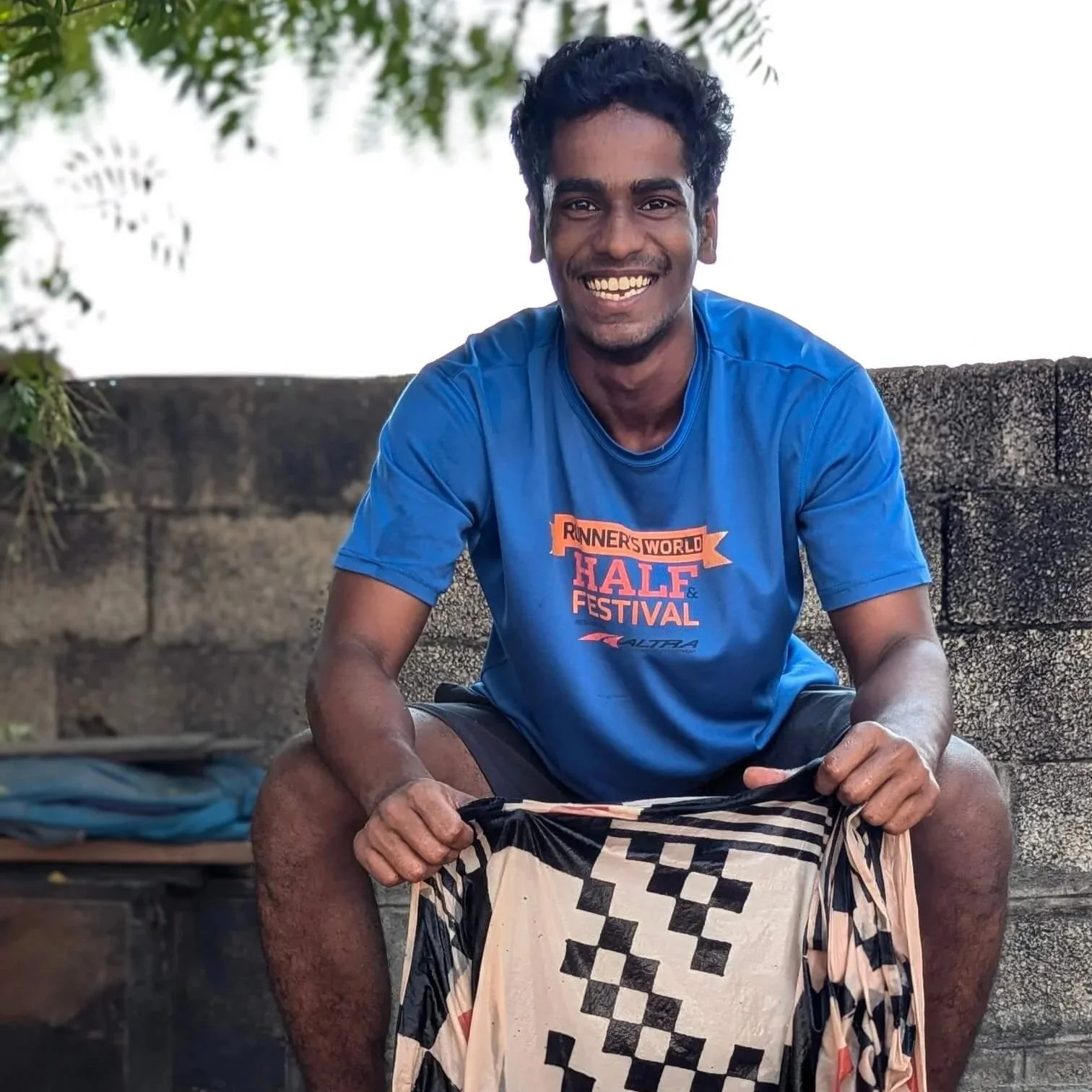 A smiling man with dark curly hair, wearing a blue t-shirt with the words "Runner's World Half & Festival" on it, sitting outdoors against a stone wall with greenery above him. He is holding a patterned fabric in his hands.