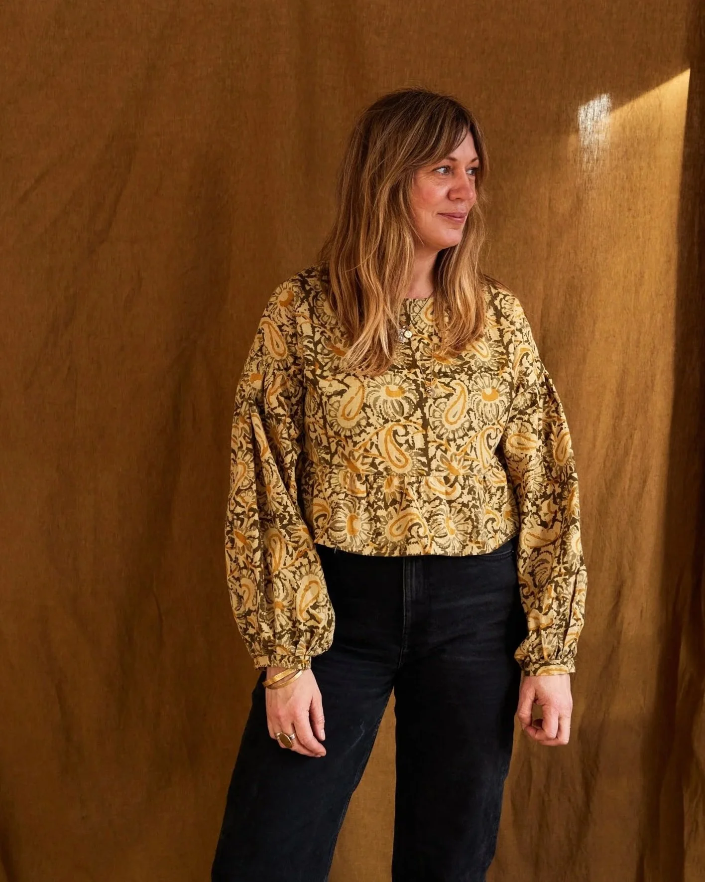 A woman with shoulder-length light brown hair wearing a beige and yellow patterned blouse and black pants standing against a brown wooden background.