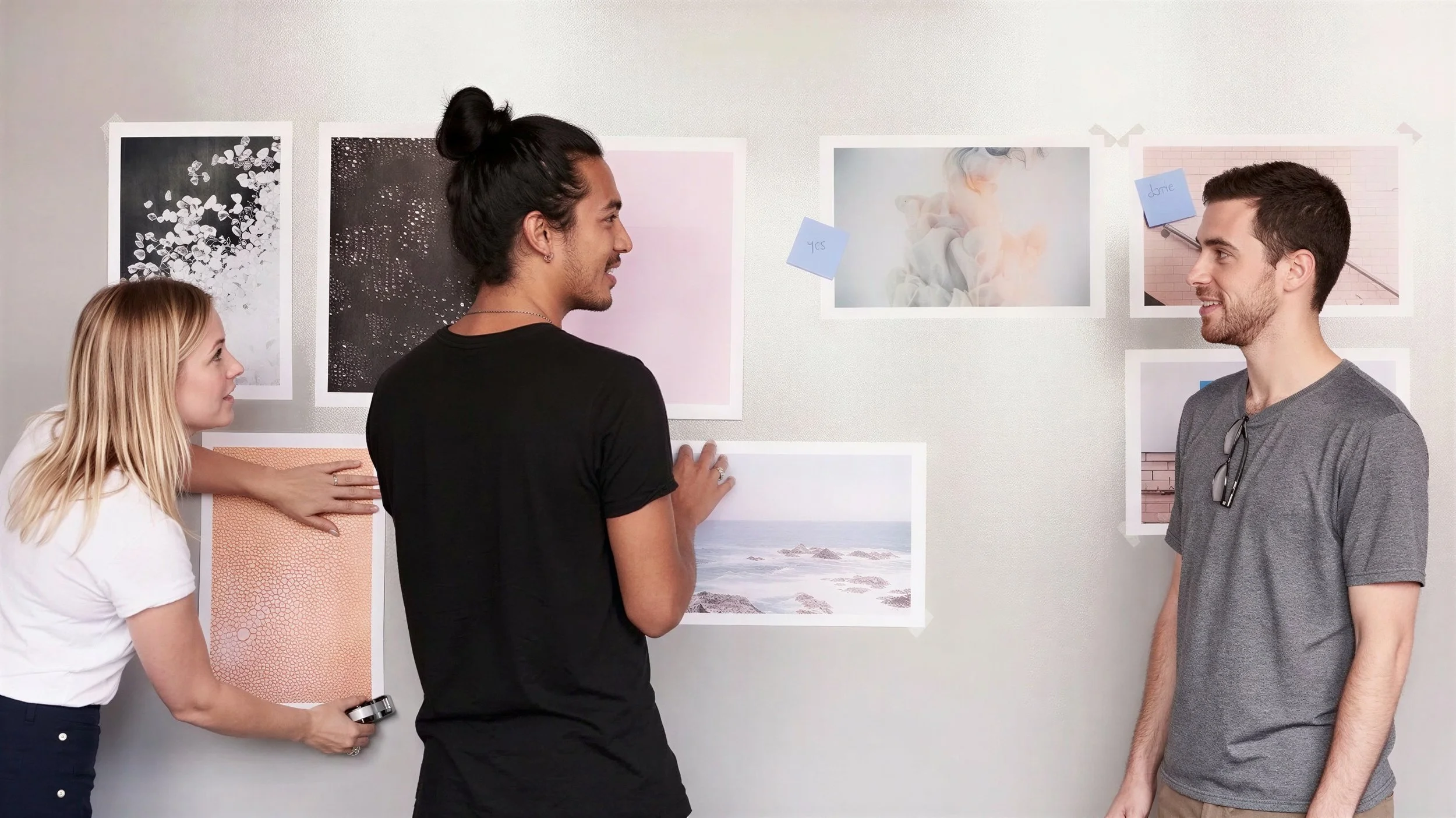 Drei junge Menschen sprechen vor einer Wand mit fotografischen Kunstwerken und Notizen, während eine Person eine Fotografie an die Wand hängt.