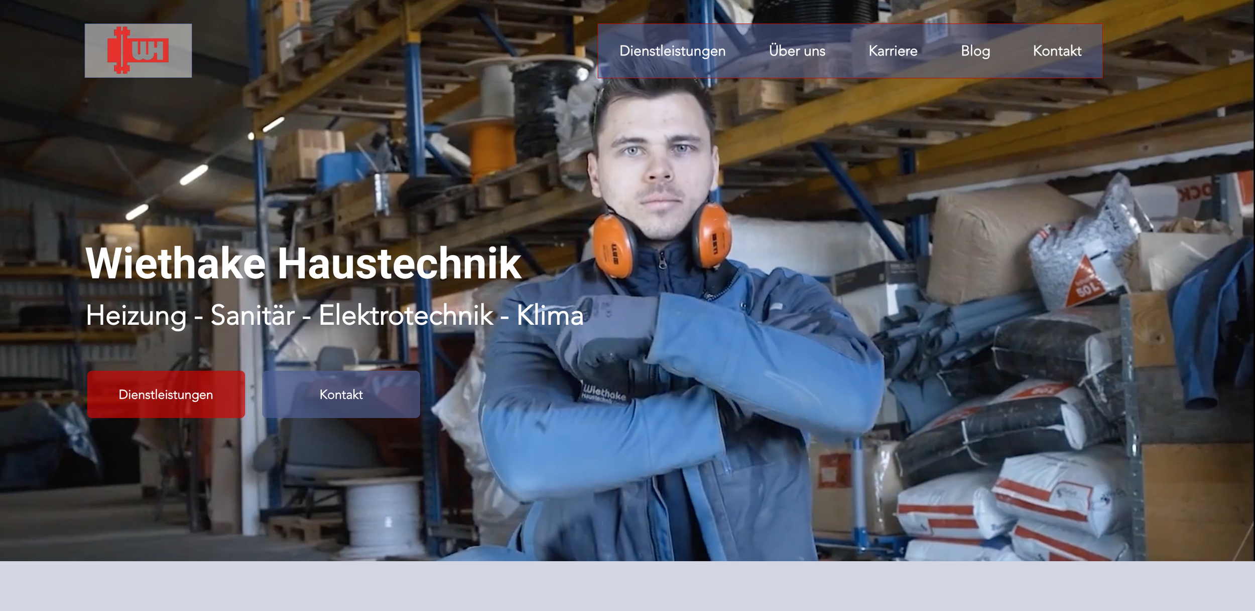 Mann mit blauer Arbeitsjacke und orange Gehörschutz im Lager mit Regalen voller Baumaterialien und Werkzeugen.