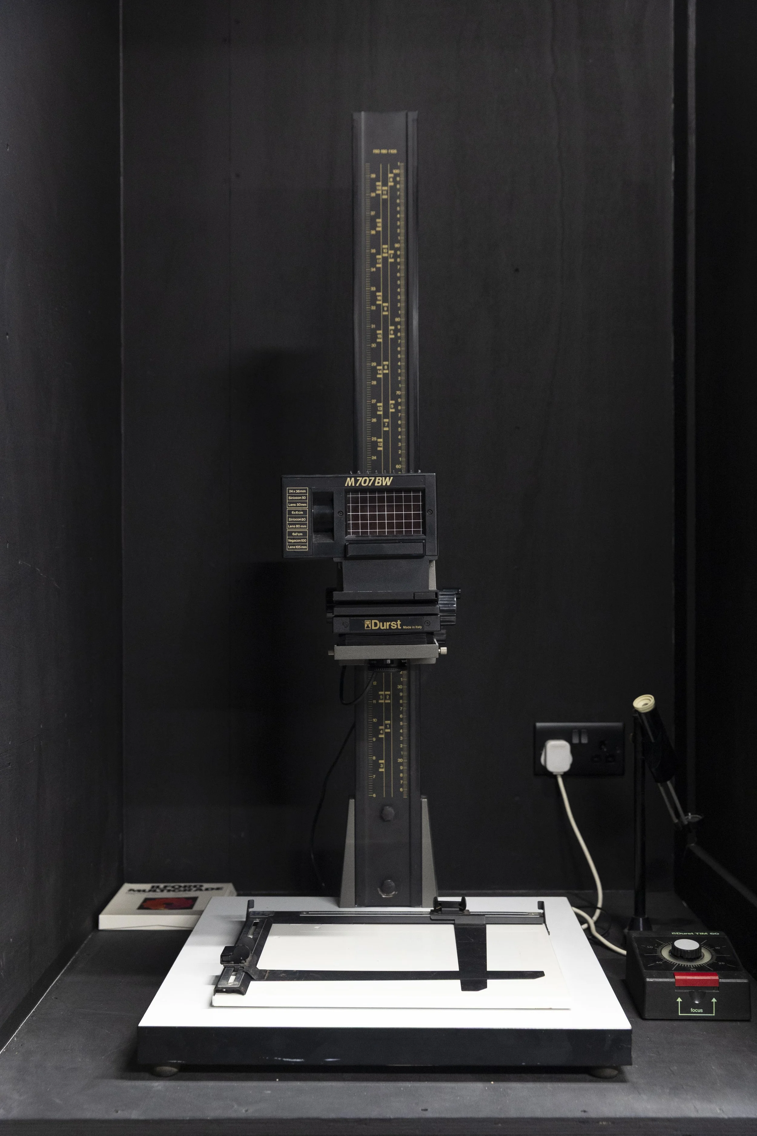A machine used for scientific or industrial purposes with a vertical part marked 'M707BW', a digital display, and a glass slide with metal clips on a flat surface