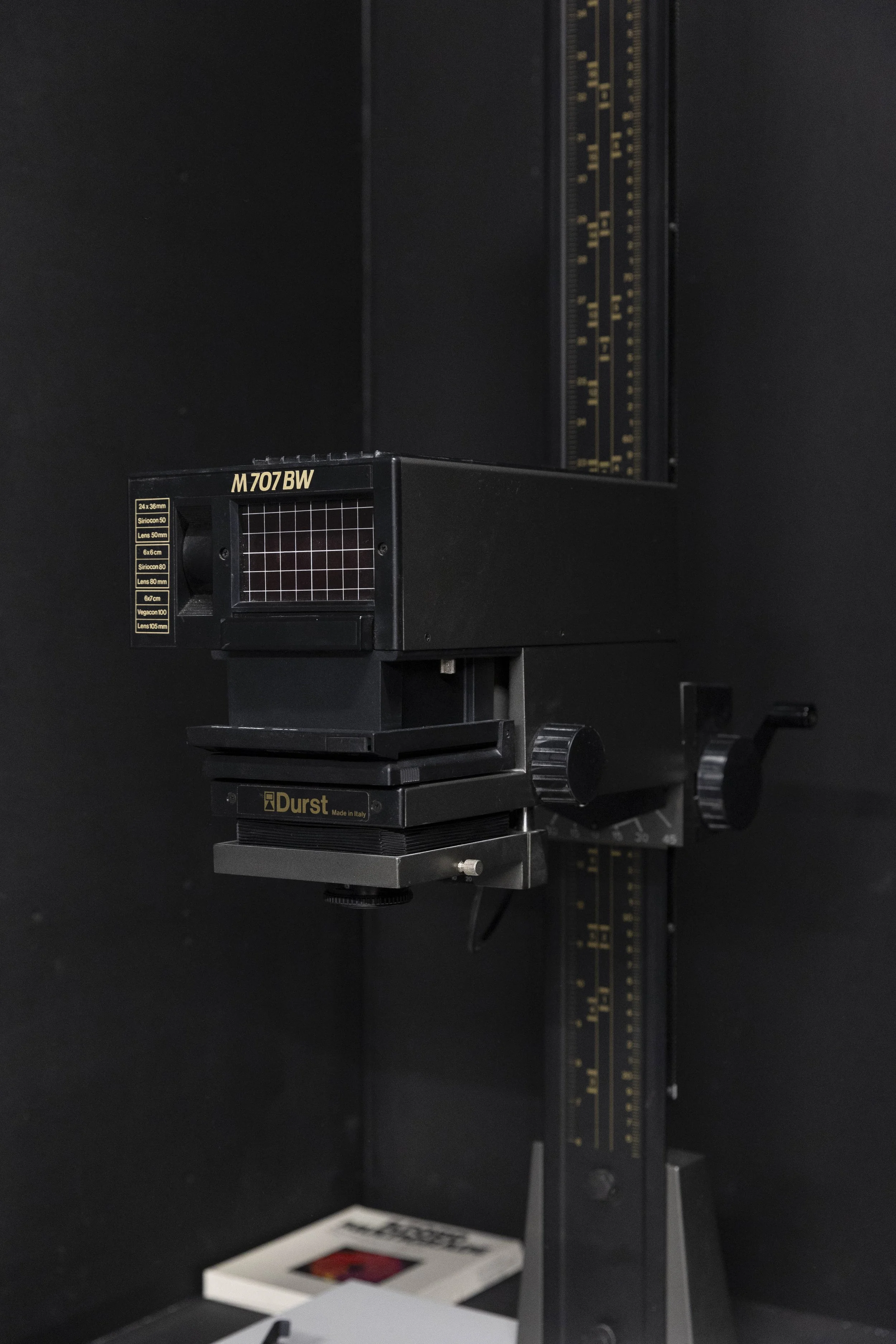 Close-up of a microscope with a black background. The microscope has a black and gold adjustment marked M707BW, and a small viewfinder. Part of a red and white object is visible at the bottom of the image.