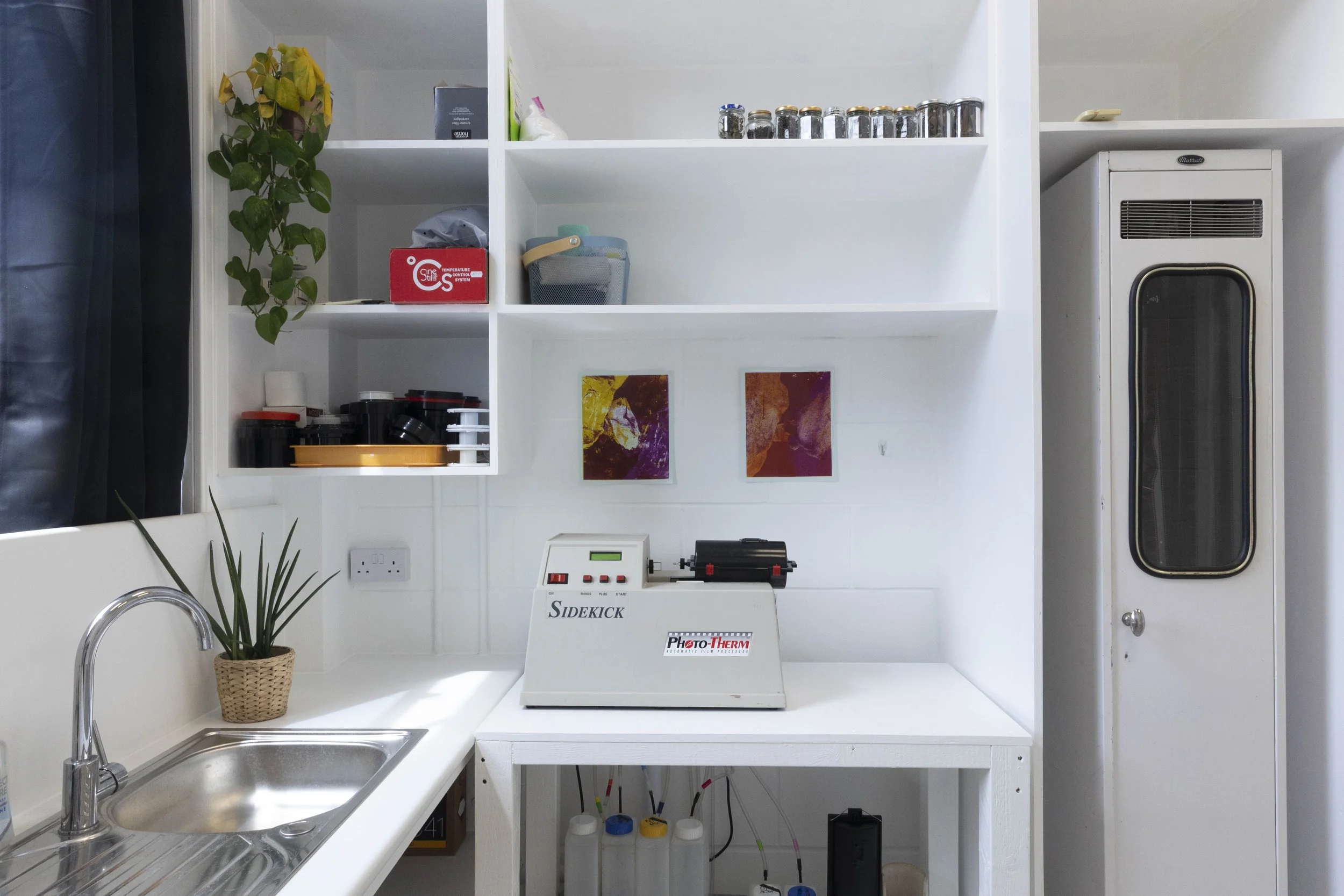 A kitchen workspace with a white countertop, stainless steel sink, potted plant, and white shelving unit with various kitchen items and a scientific instrument labeled Sidekick and Photo-Therm.