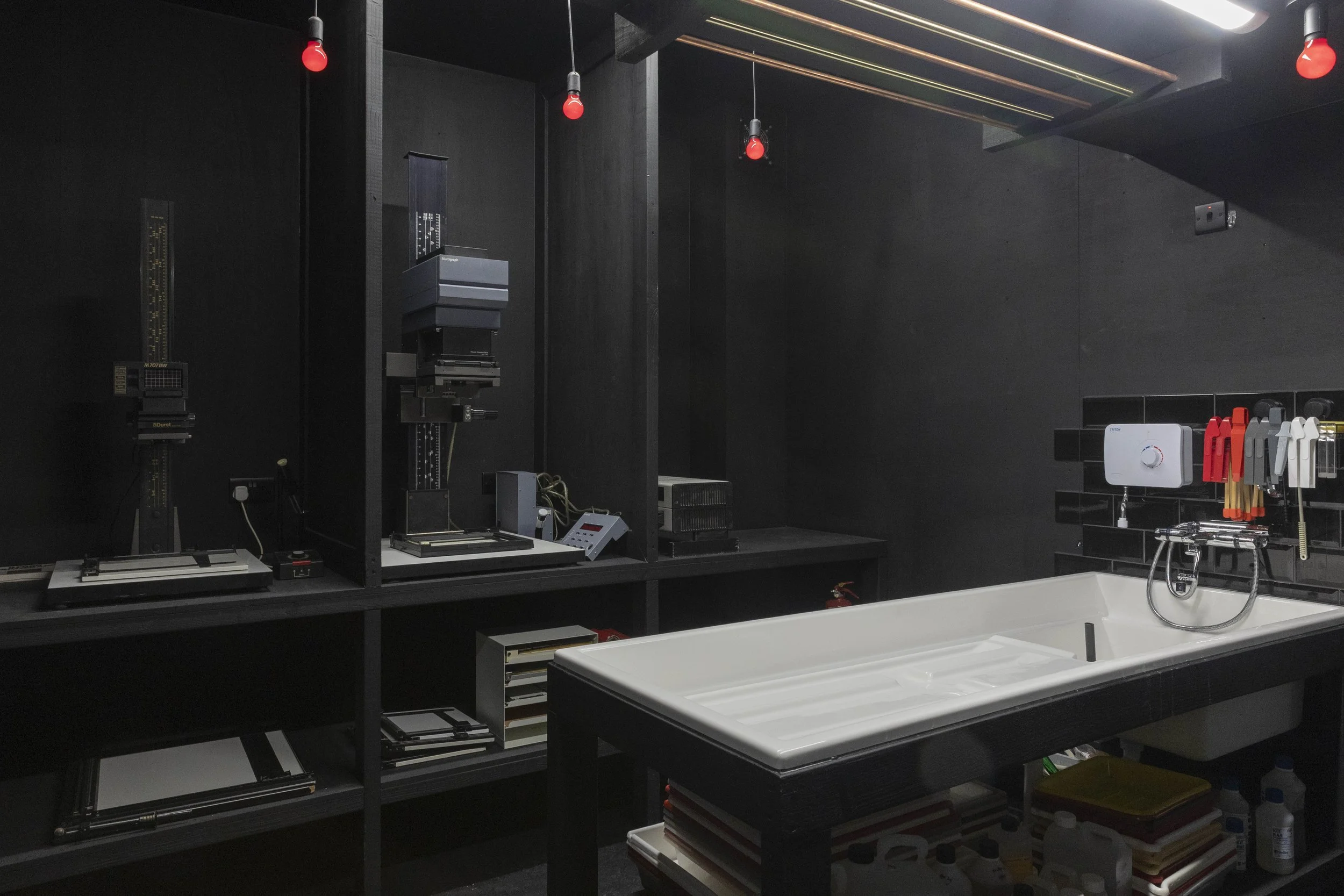 A laboratory workspace with black walls and shelves containing scientific instruments, tools, and supplies. A white sink or basin is visible in the lower right corner with various laboratory equipment and red, gray, and white gloves hanging on hooks above the sink.