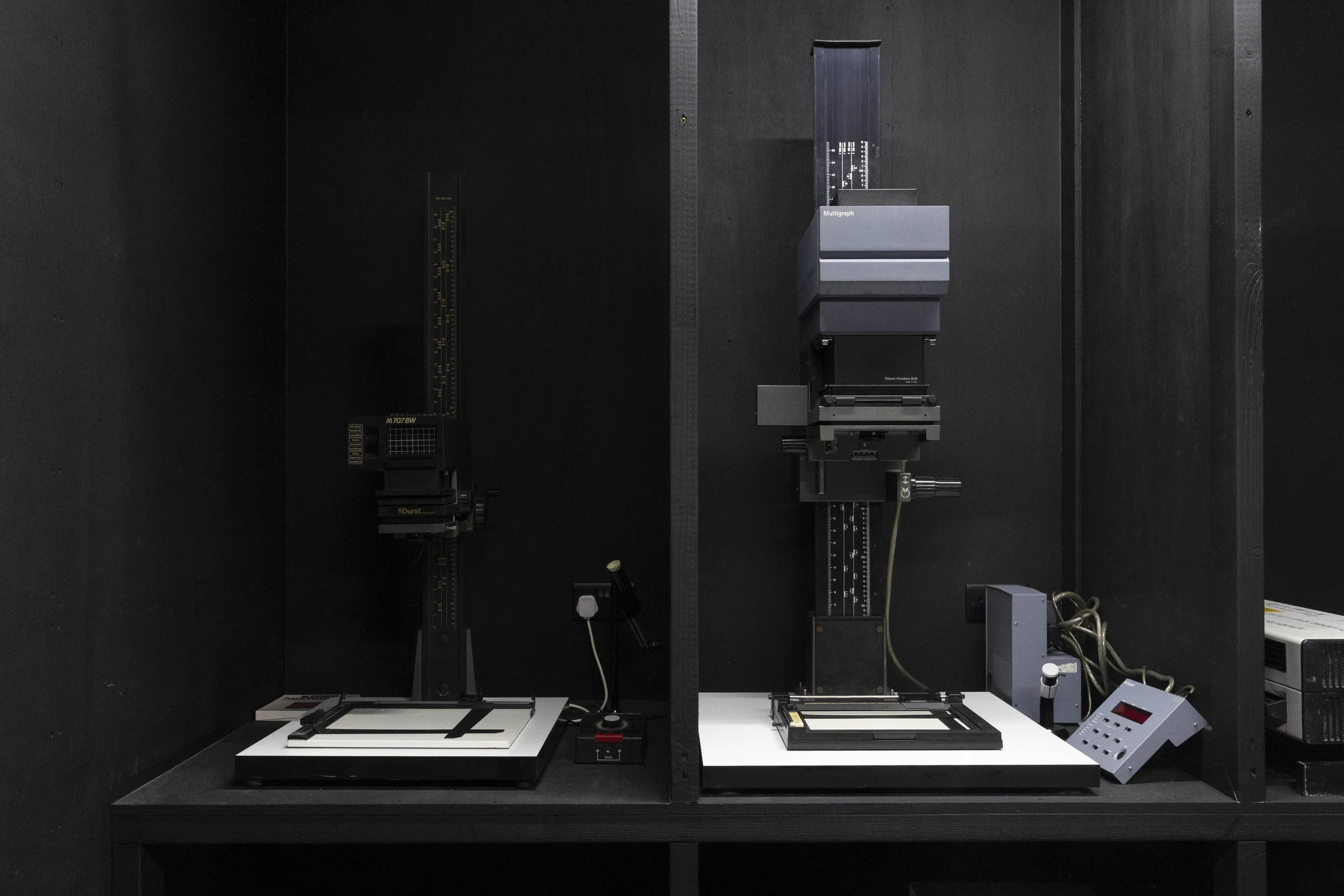 Laboratory setup with two optical breadboards, various mechanical components, and measurement devices mounted on a black wall background.