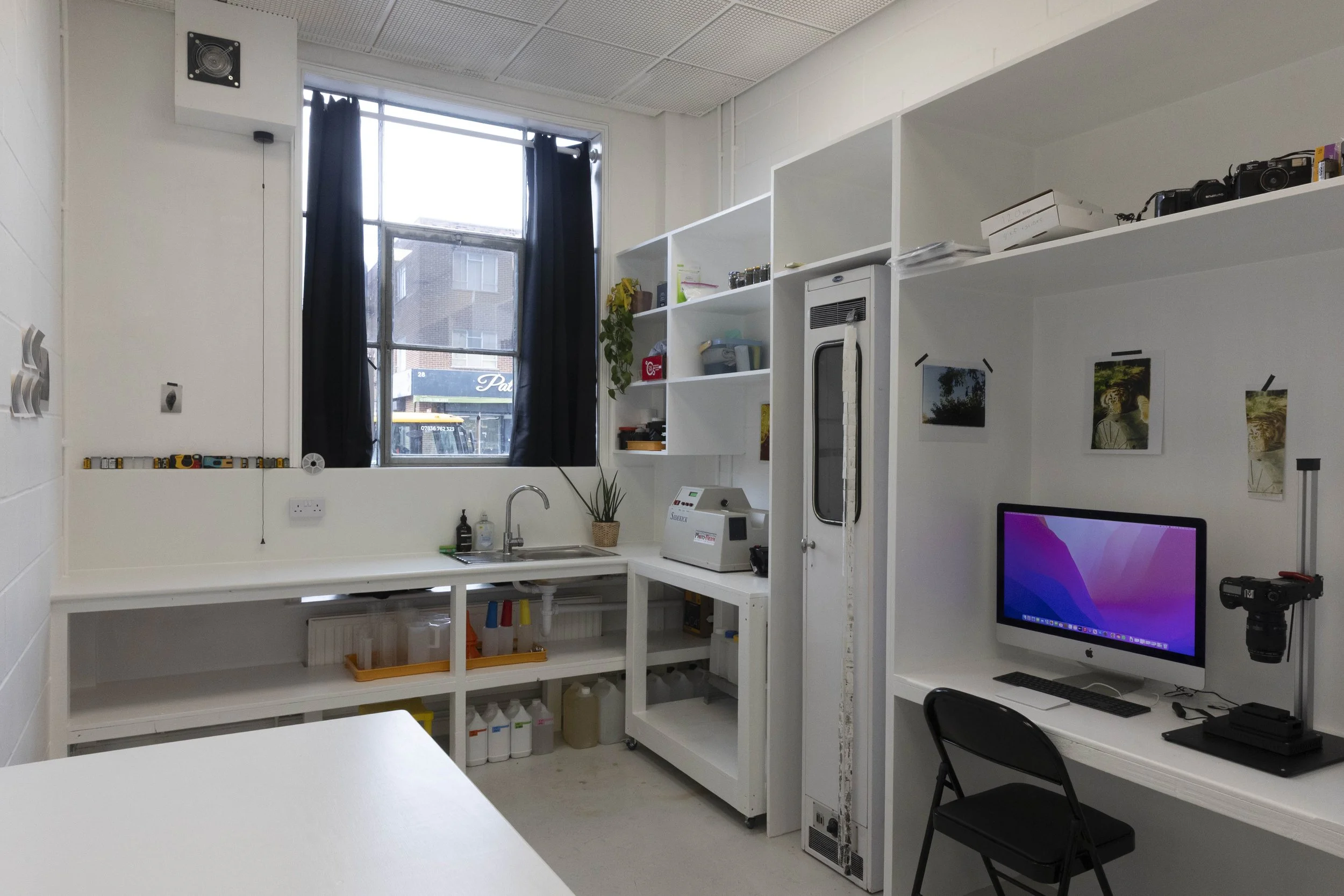 A room with white walls and a large window covered with dark curtains. There is a white shelving unit with various storage boxes, plants, and equipment. A computer monitor, keyboard, and camera are visible on a desk in the corner.