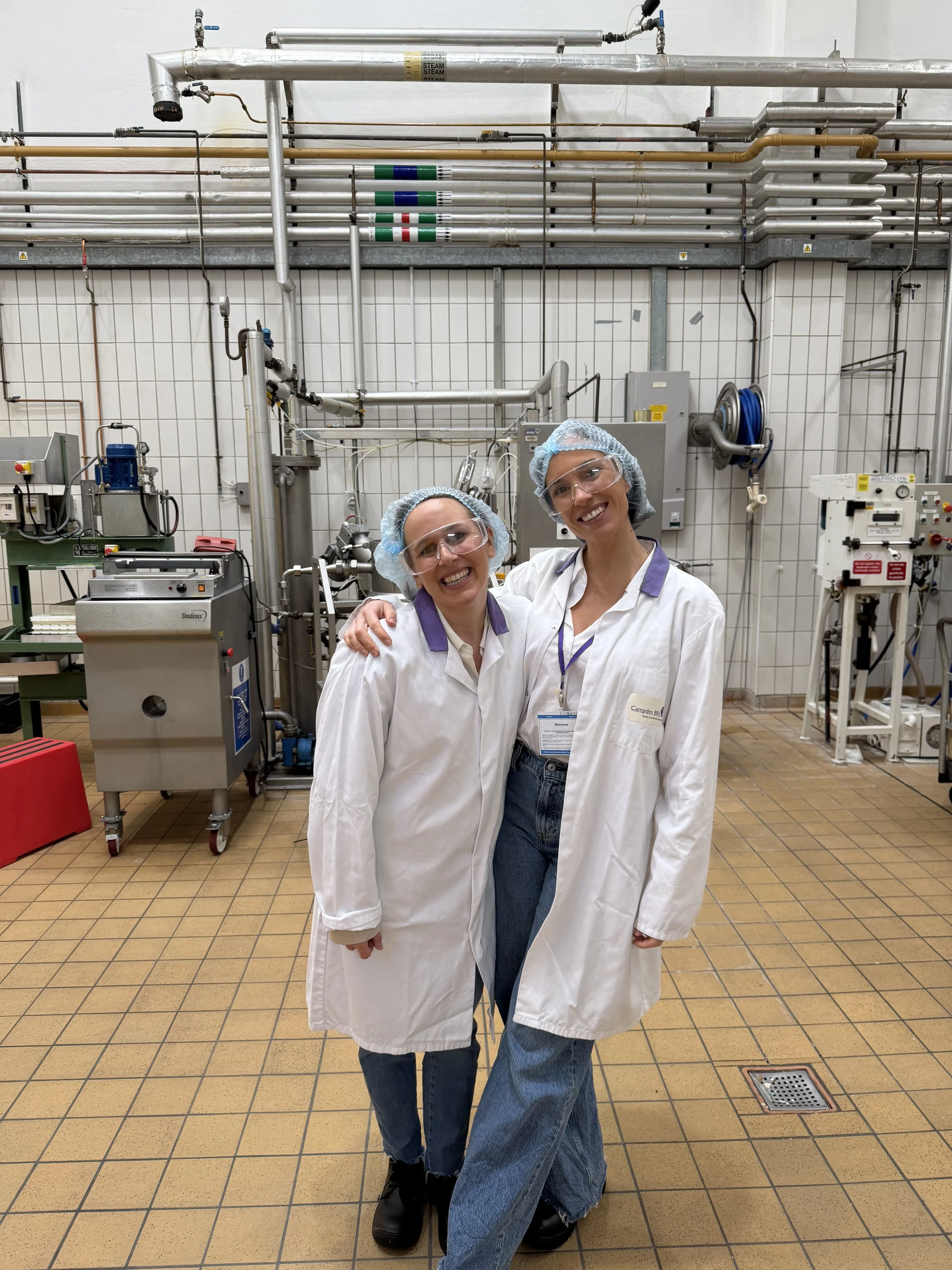 Two smiling women wearing lab coats, safety glasses, and hairnets in a laboratory or industrial setting with pipes, tanks, and equipment behind them.