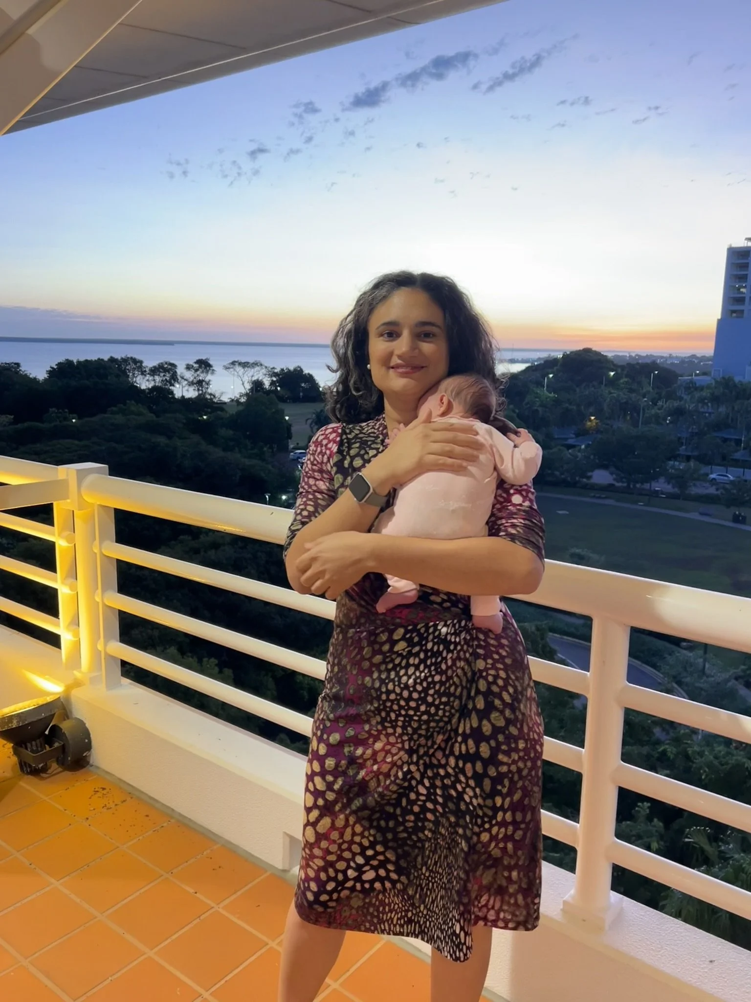 A woman holding a baby on a balcony at the Northern Territory's Parliament House during sunset, with a view of trees, water, and a cityscape in the background.