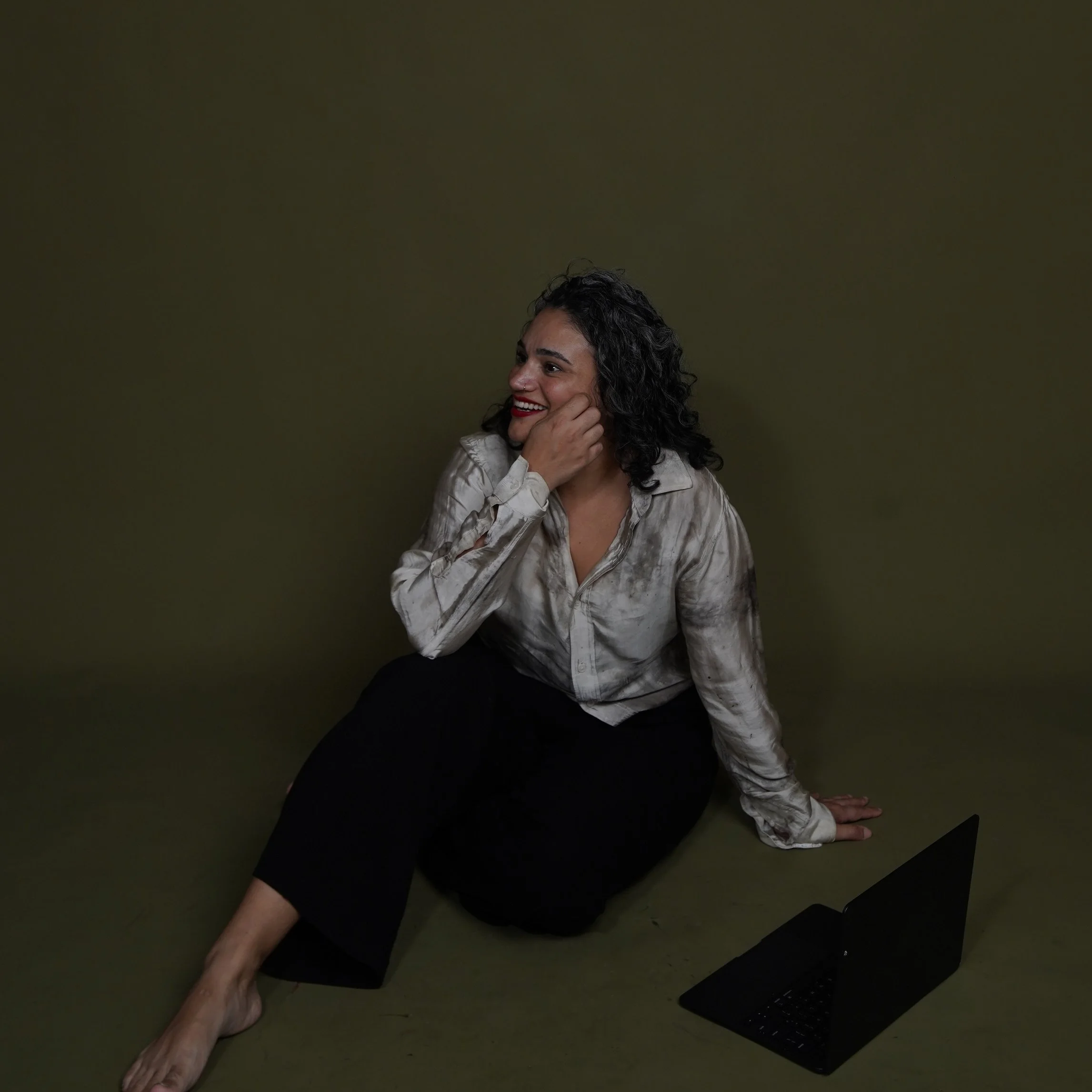 A woman with dark, curly hair, wearing red lipstick, a white button-up shirt, and black pants, is sitting on the floor against a dark green wall, smiling and resting her head on her hand, with a black computer keyboard on the floor nearby.