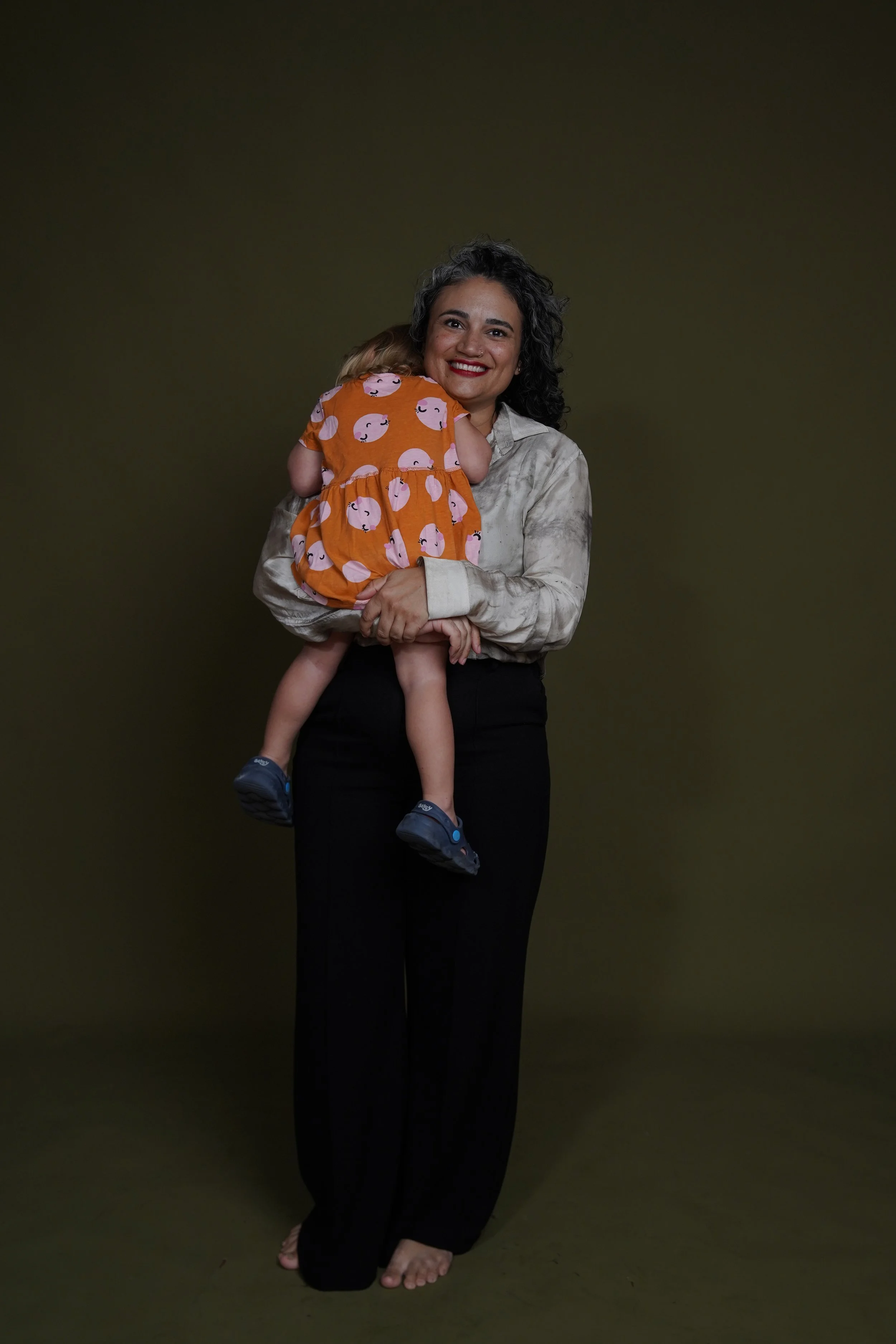 Lauren with curly hair, smiling with red lipstick, standing barefoot and holding a young child. The child, facing her, has blonde hair, and is wearing an orange dress with a pink cartoon face pattern. The background is plain dark green.