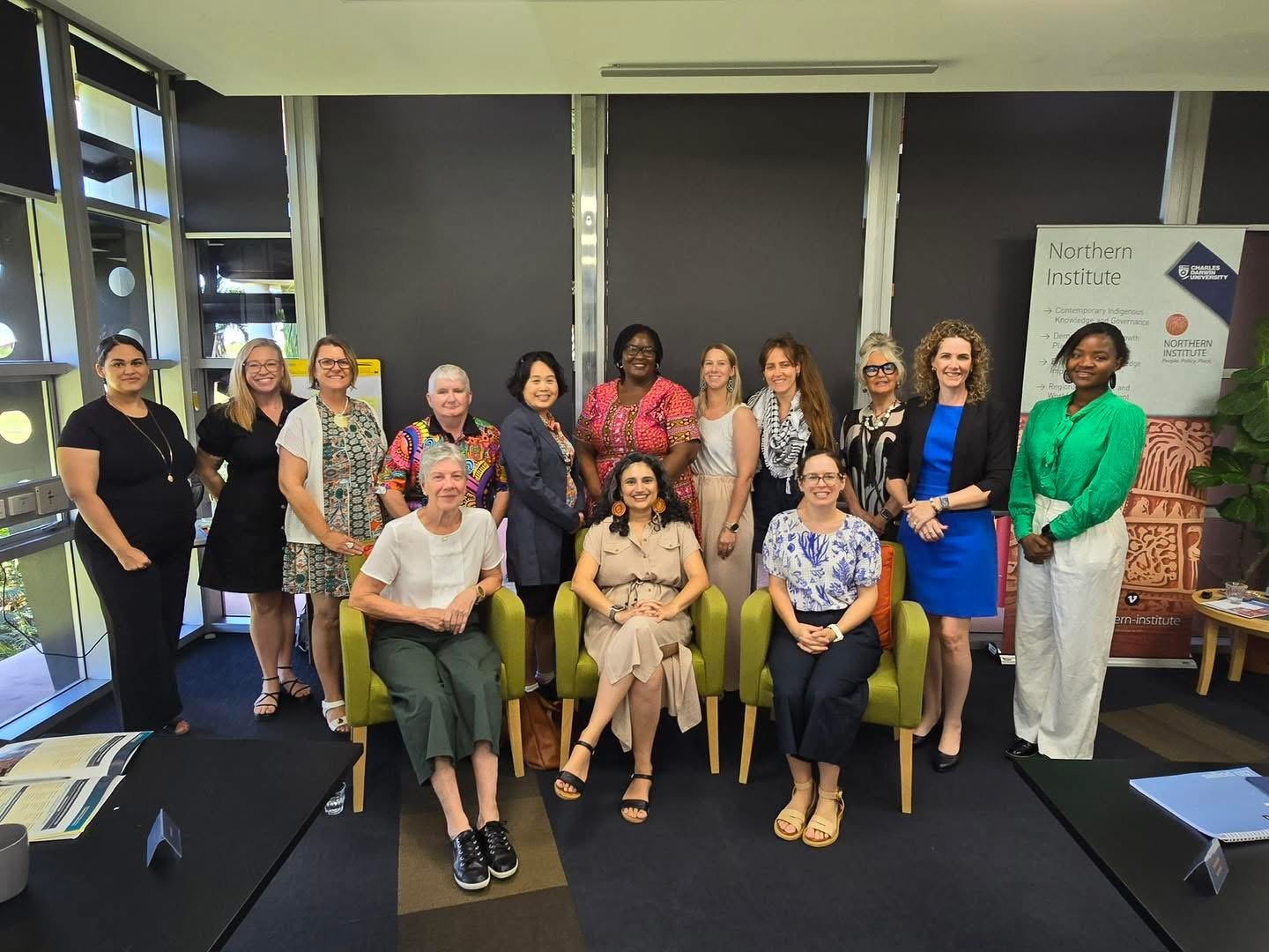 Group of diverse women smiling and posing for a photo in a conference room with some seated in front and others standing behind. There is a Northern Institute poster and a plant in the background.