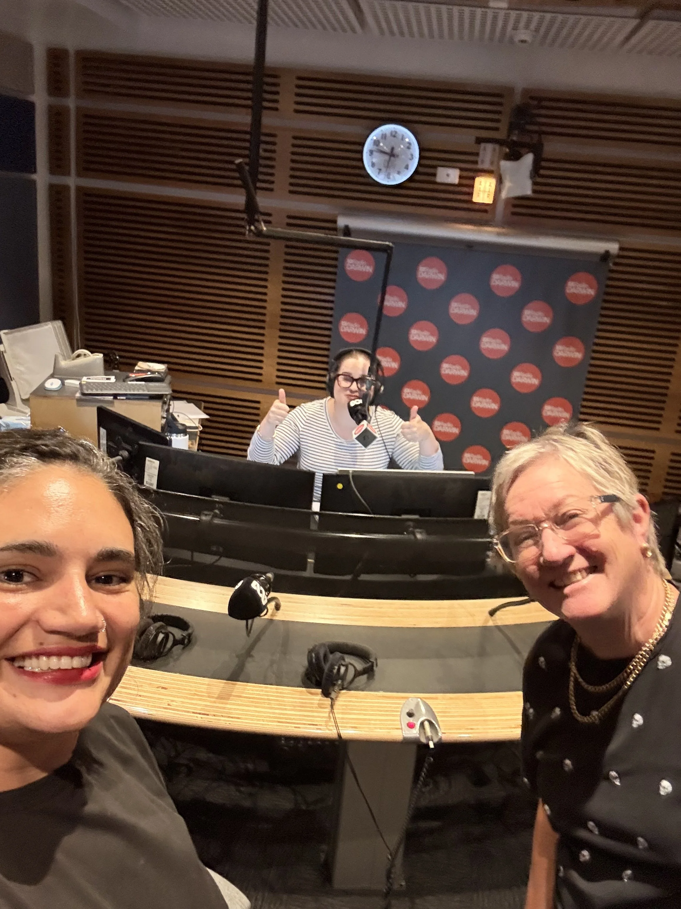 A group of people in a radio studio. Two women at the forefront are smiling and taking a selfie, while a woman in the background is speaking into a microphone. The studio has wood-paneled walls, a clock, and various broadcasting equipment.