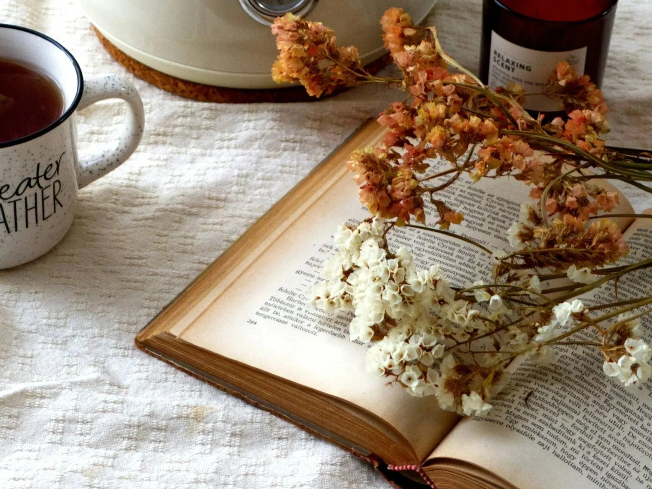 Libro abierto con flores secas sobre él, taza con bebida y frascos en un fondo de tela beige.