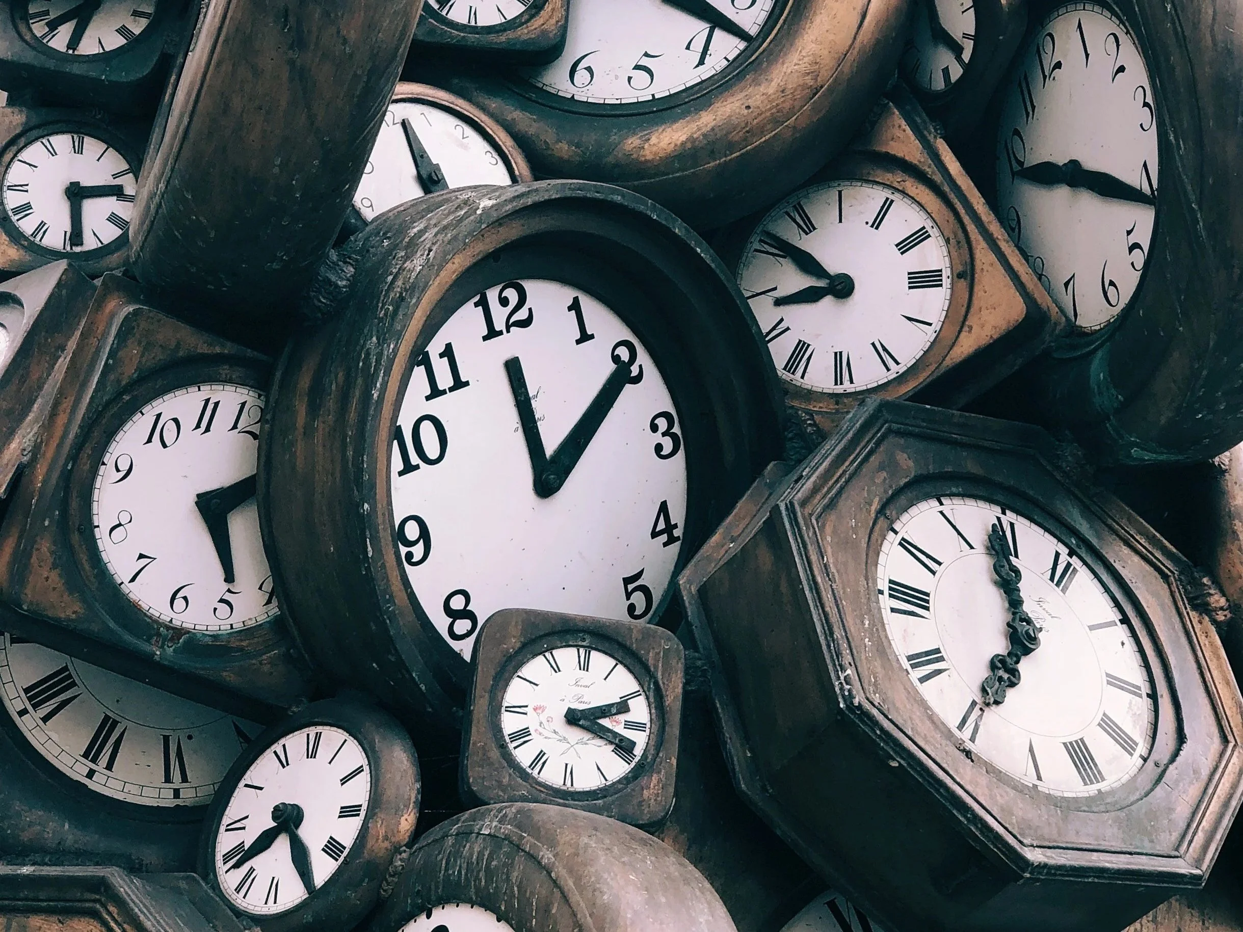 Varios relojes de pared antiguos apilados, algunos con caras cuadradas y otros redondas, mostrando diferentes horas.