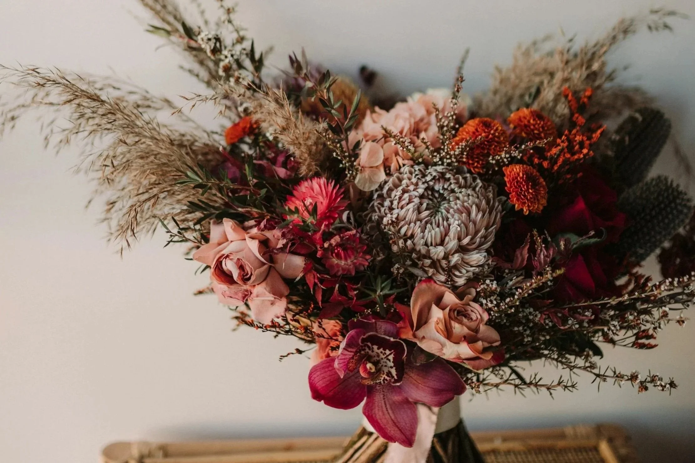 Ramo de flores con variedad de flores en tonos rosados, rojos y naranjas, incluyendo orquídeas, crisantemos y flores de hierba seca, sobre fondo neutro.