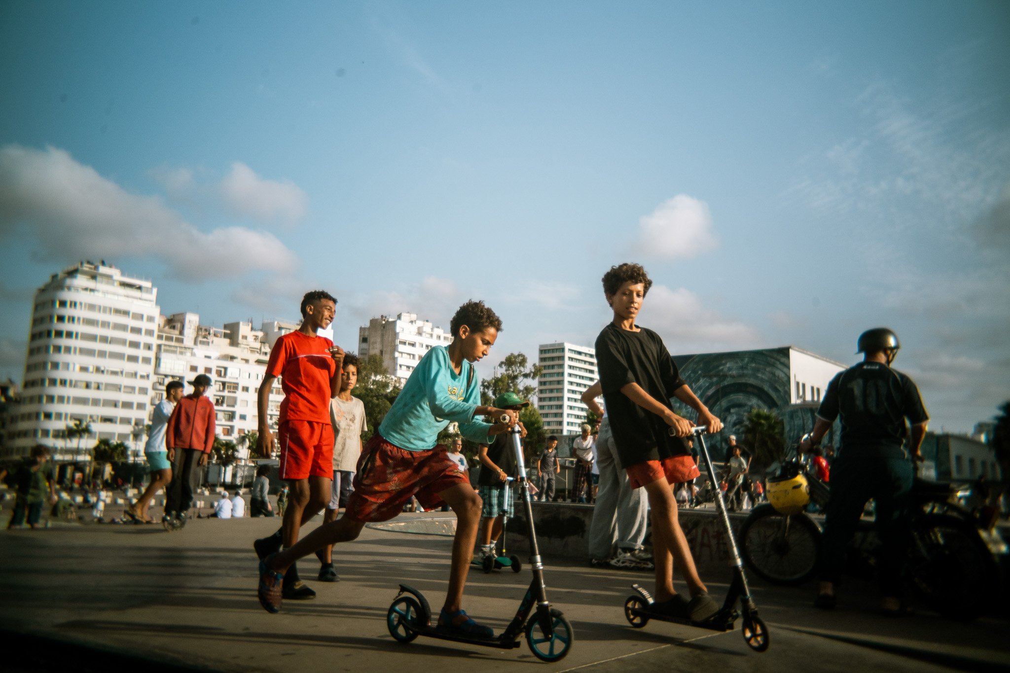 Young people riding scooters and bicycles at an outdoor skate park with city buildings in the background