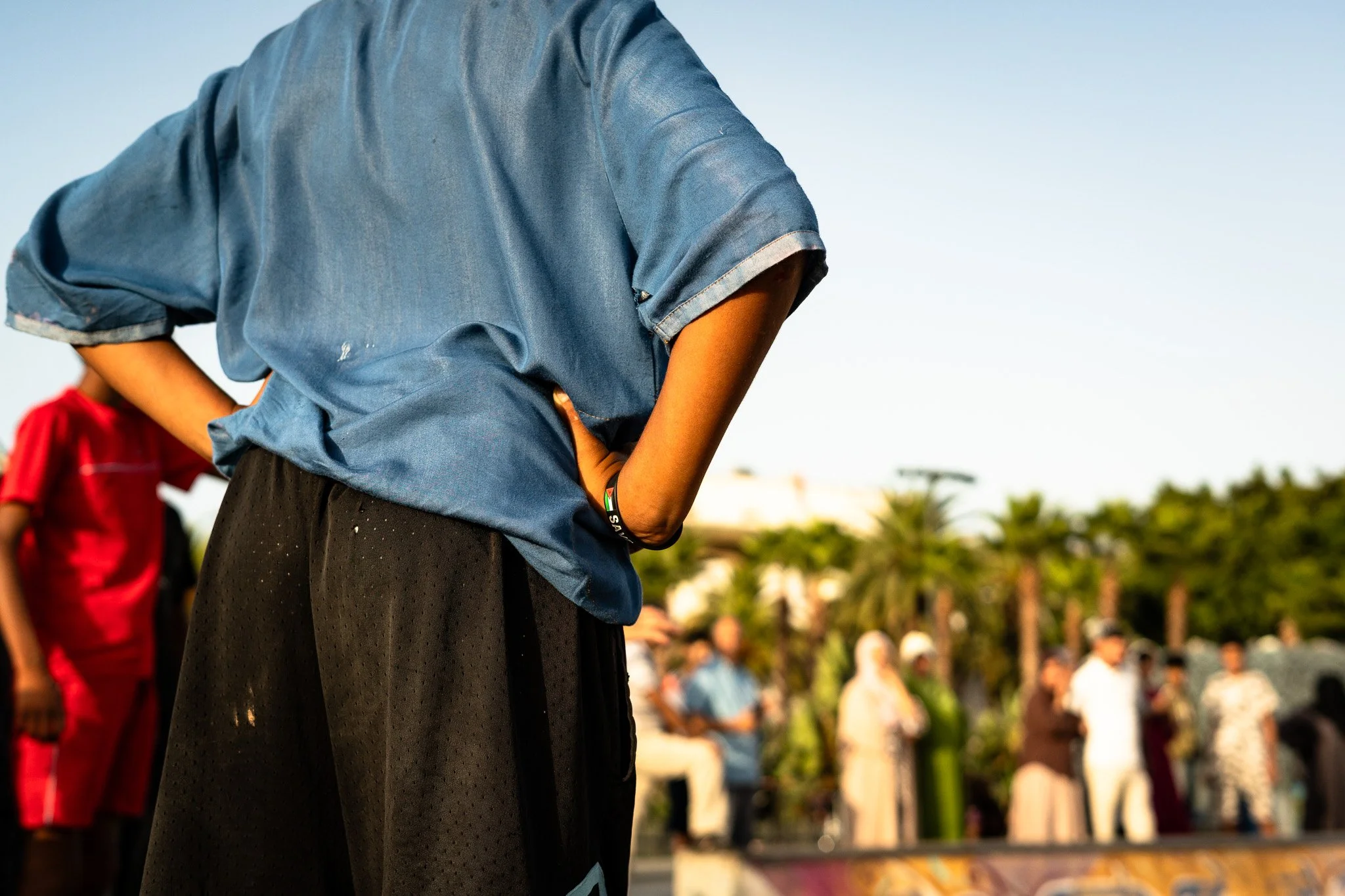Person in a blue shirt and black skirt standing outdoors with hands on hips, with a blurred crowd and palm trees in the background