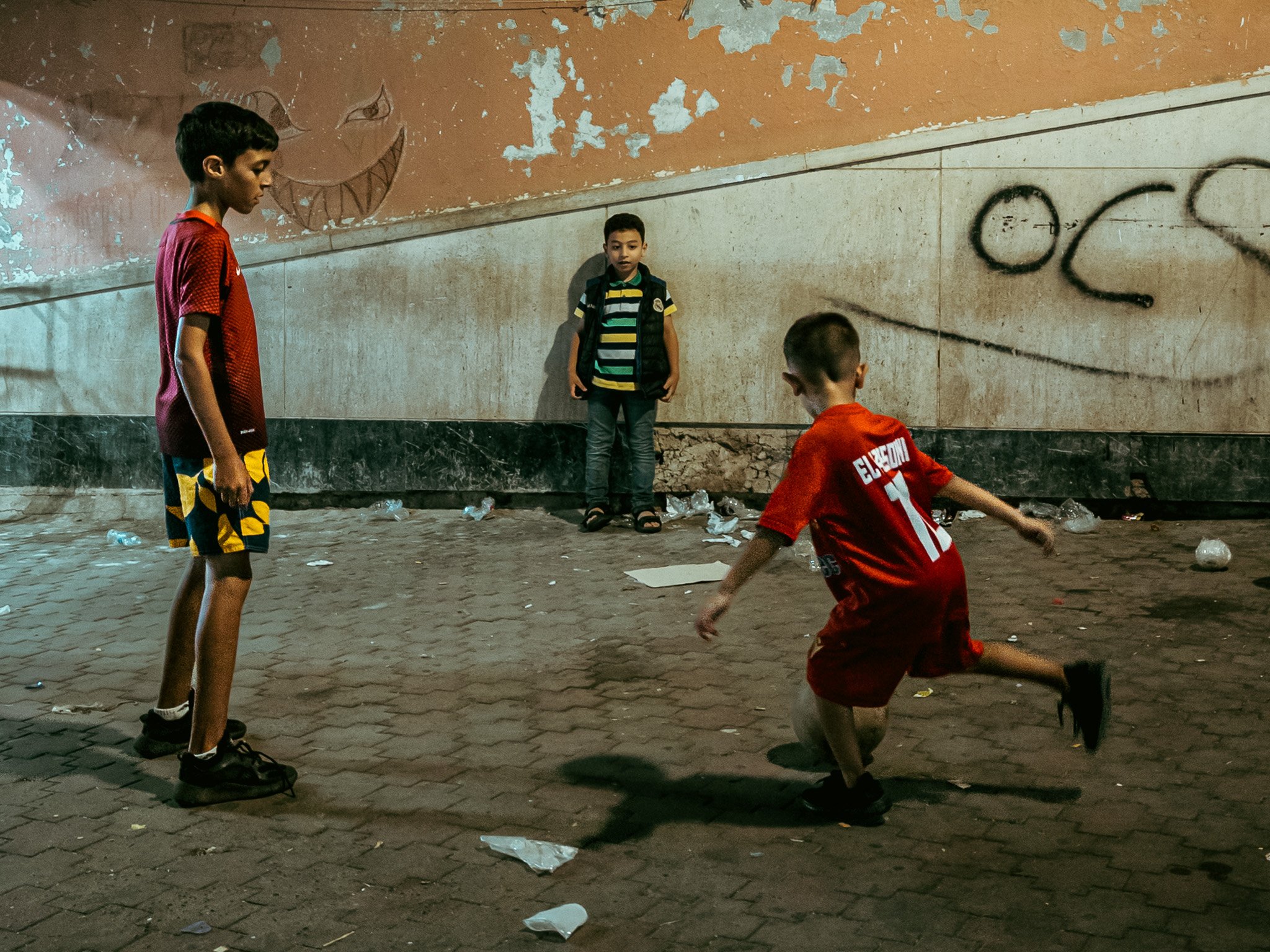 Four boys are playing on a street at night, surrounded by crumpled paper and trash. Two of the boys are standing, with one in red sports clothes and the other in yellow sports clothes, looking at the boy in red playing on the ground. The third boy, i