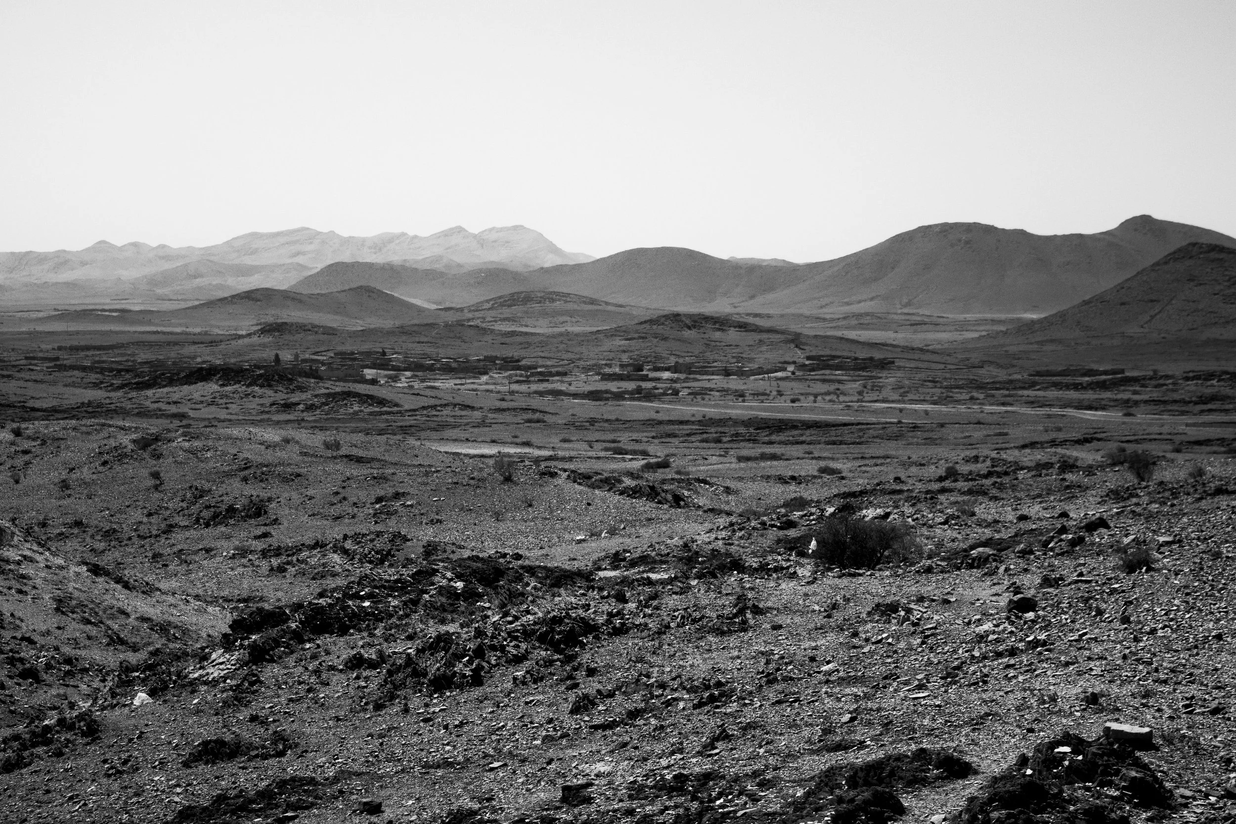 Black and white photograph of a desert landscape with scattered rocks and sparse vegetation in the foreground, rolling hills in the middle ground, and distant mountains under a clear sky in the background.