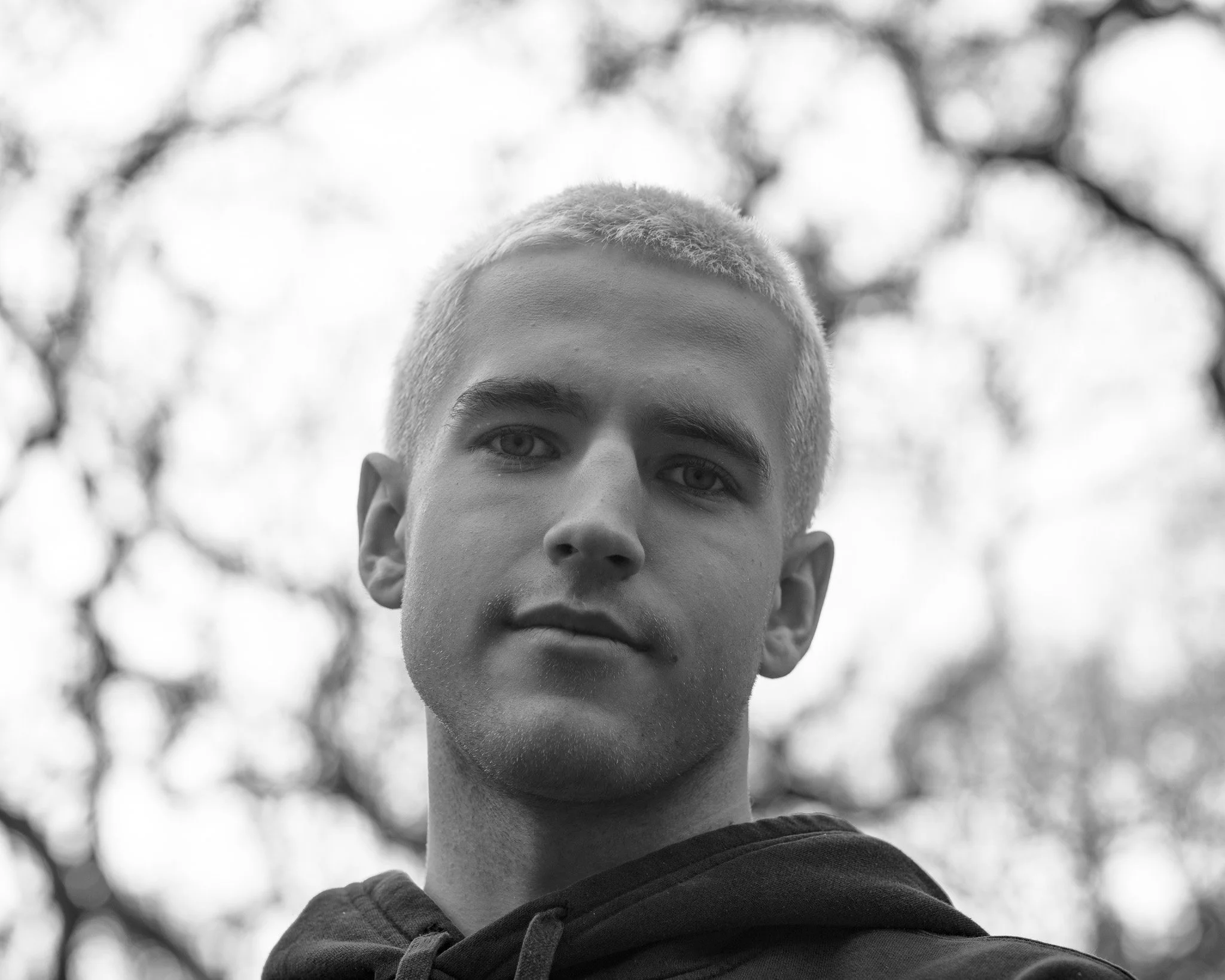 Close-up black-and-white photo of young man's face, focusing on eyes, nose, and hairstyle, with blurred tree branches in background.