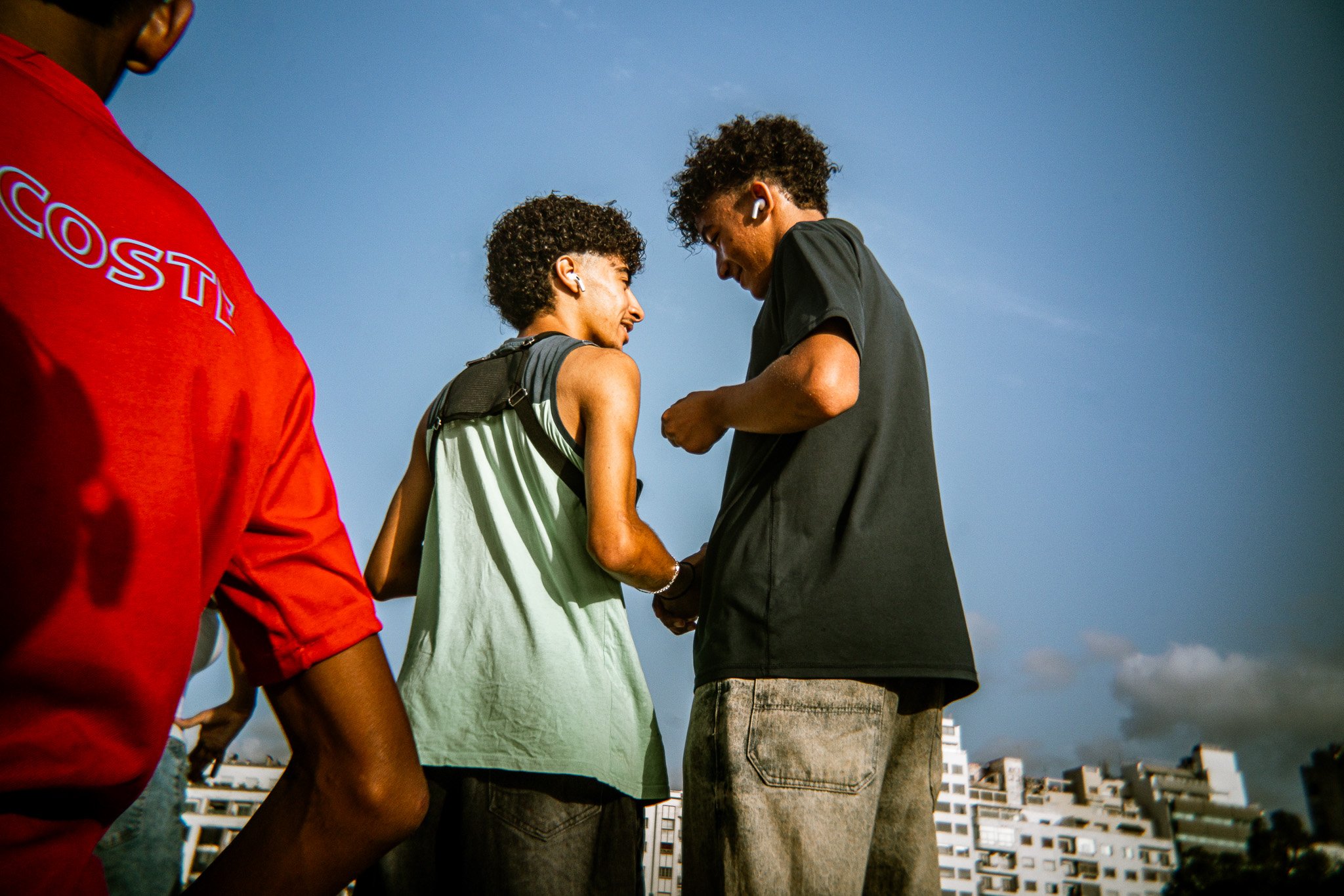 Three young men wearing casual clothing and wireless earbuds outdoors against a blue sky and city skyline