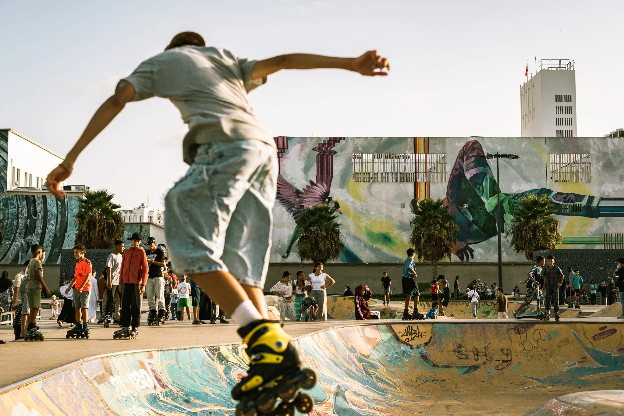 Skater performing a trick in a crowded skatepark with palm trees and colorful murals.