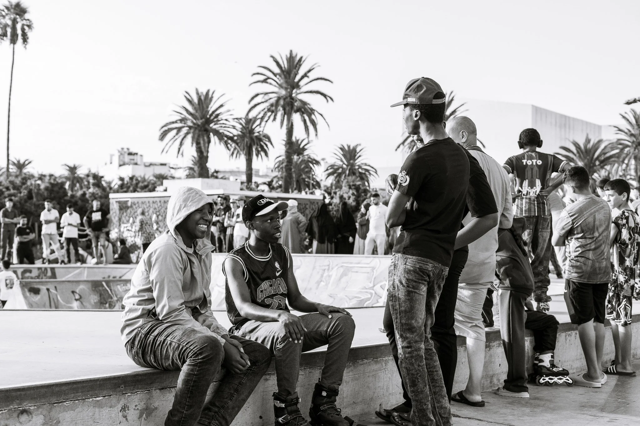 People socializing at a palm-lined outdoor skatepark, with skaters and spectators gathered around graffiti-covered ramps.