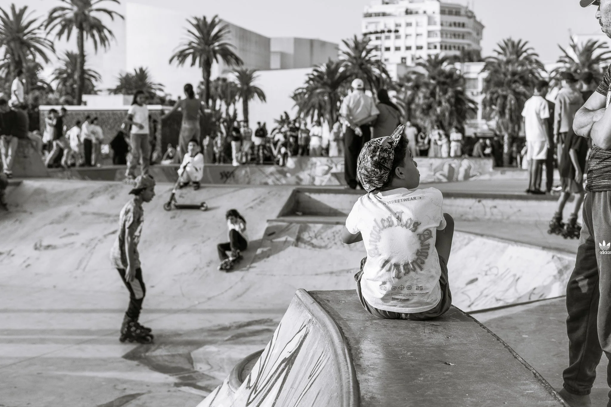 Black-and-white photo of children and adults at a palm-lined outdoor skate park, with a child sitting on a concrete ramp in the foreground.