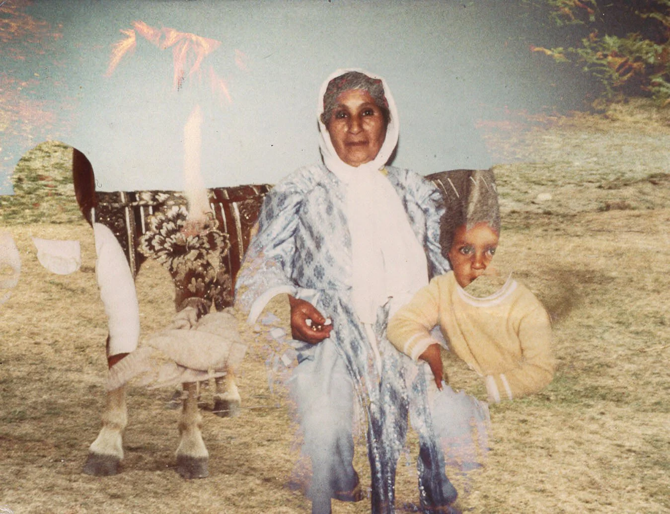 An elderly woman in traditional clothing and a young boy stand outdoors in front of a painted backdrop of landscape scenery. The woman is seated on a decorated chair, and the boy is standing next to her with his hand on his hip.